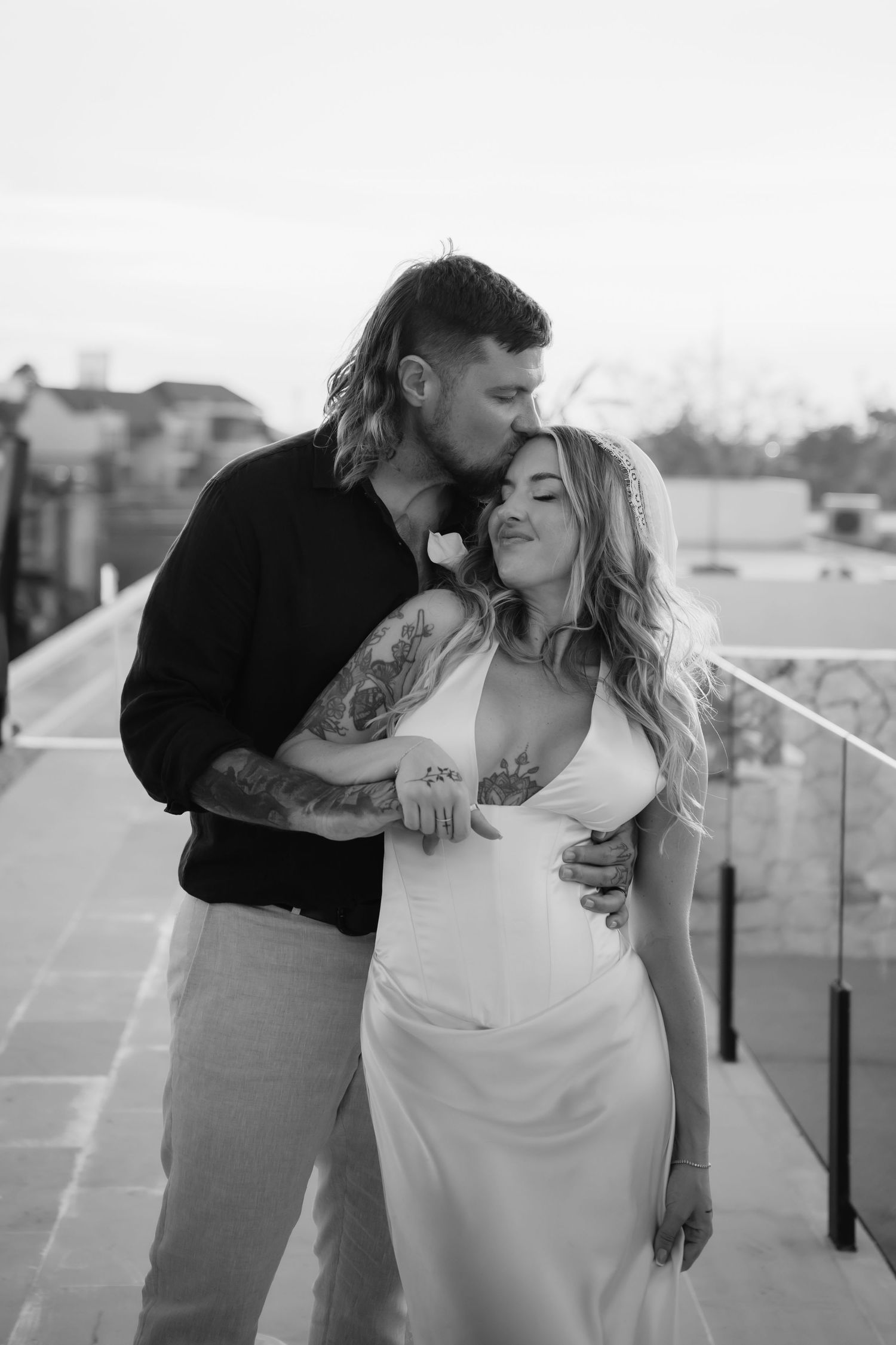 Black and white photo of tattooed groom kissing bride on cheek during rooftop wedding portrait.