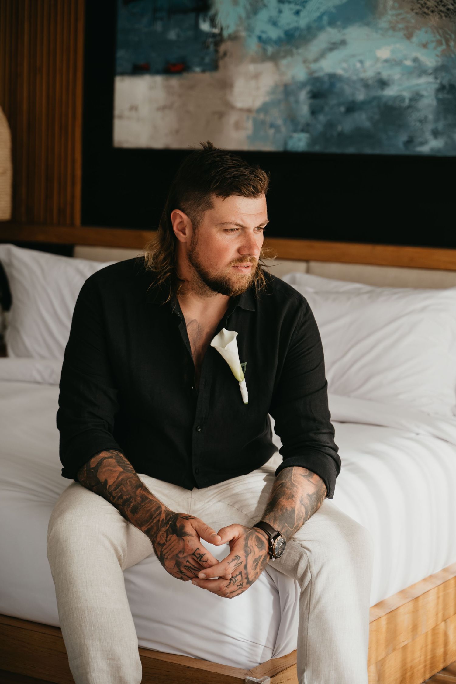 Tattooed groom in black shirt and white pants sits on bed with boutonniere, looking away thoughtfully.