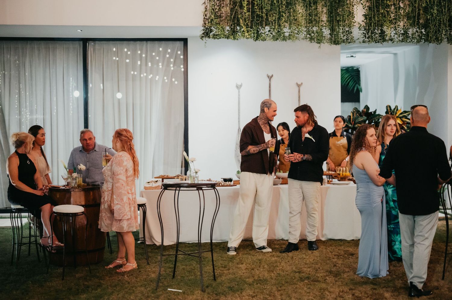 Wedding guests mingle and socialize at an elegant indoor reception with greenery wall and string lights.