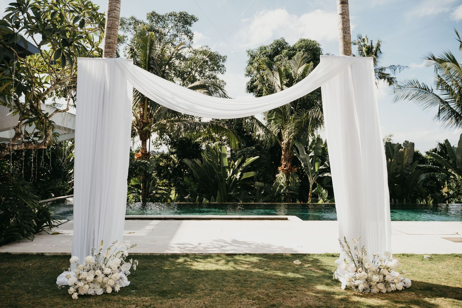 Elegant outdoor wedding arch draped in white fabric with floral arrangements, set against a tropical pool backdrop.