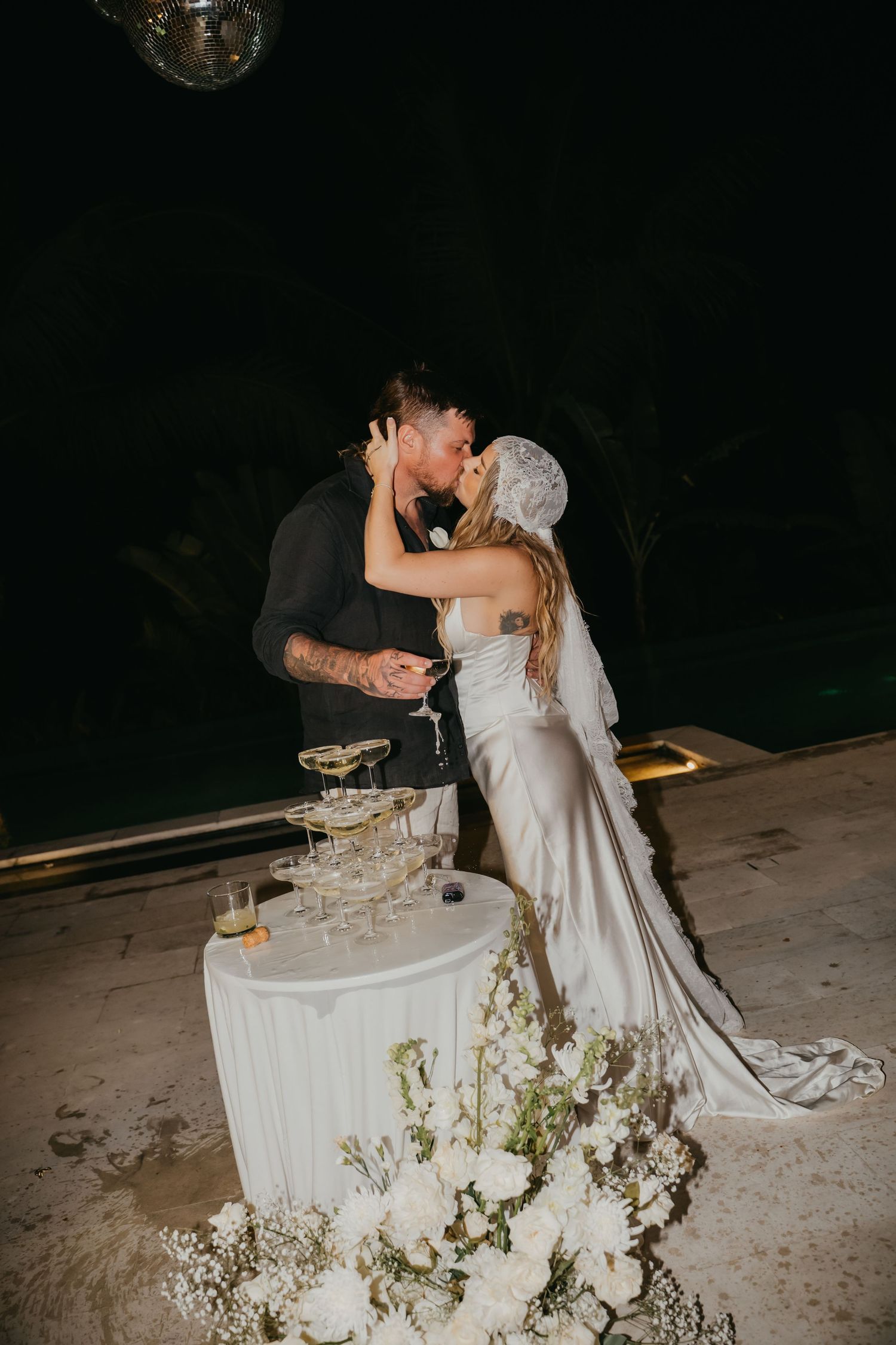 Bride and groom share a kiss beside a champagne tower with white floral arrangements at an outdoor nighttime wedding reception.