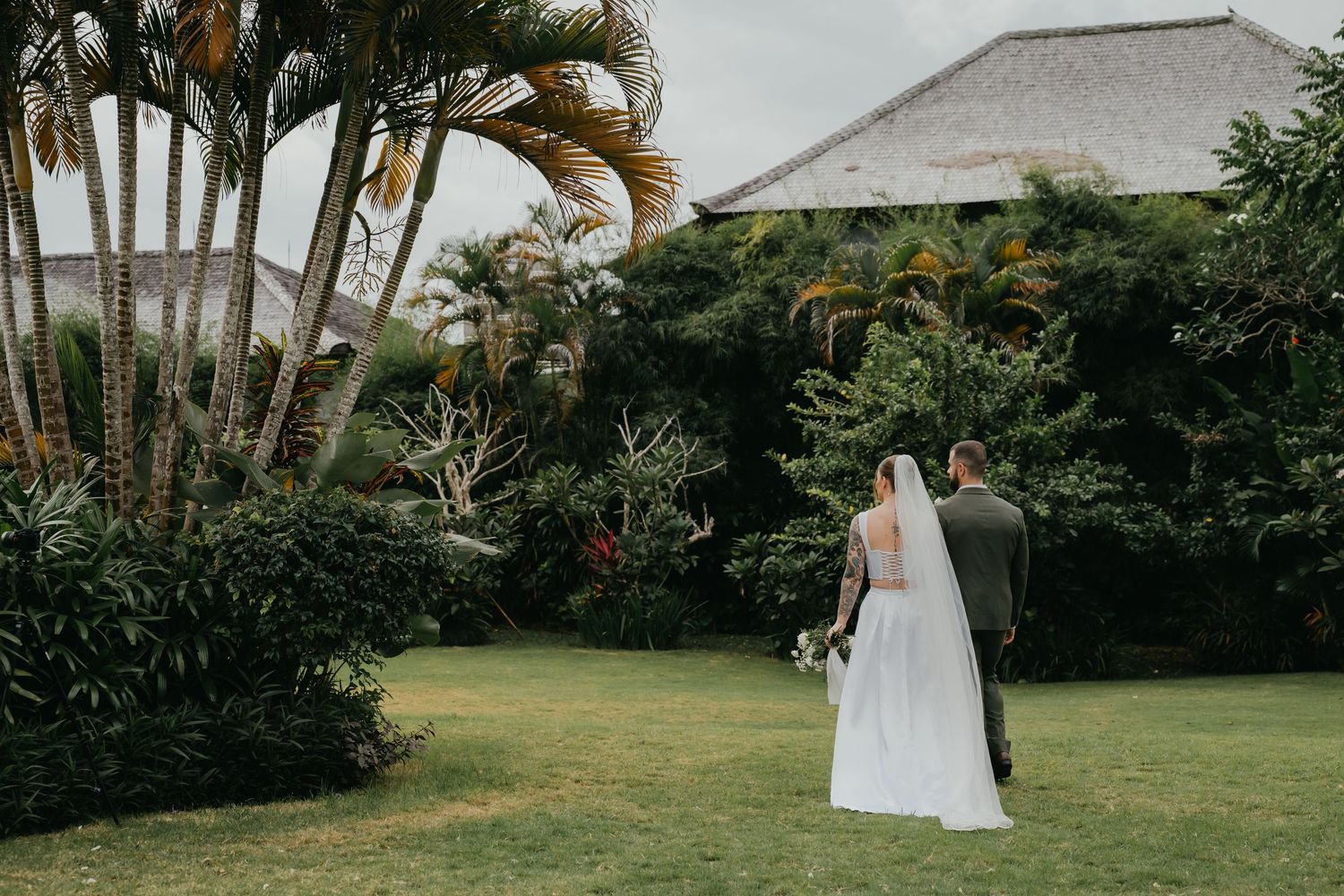 Bride and groom walk hand in hand through lush tropical garden surrounded by palm trees and green foliage.