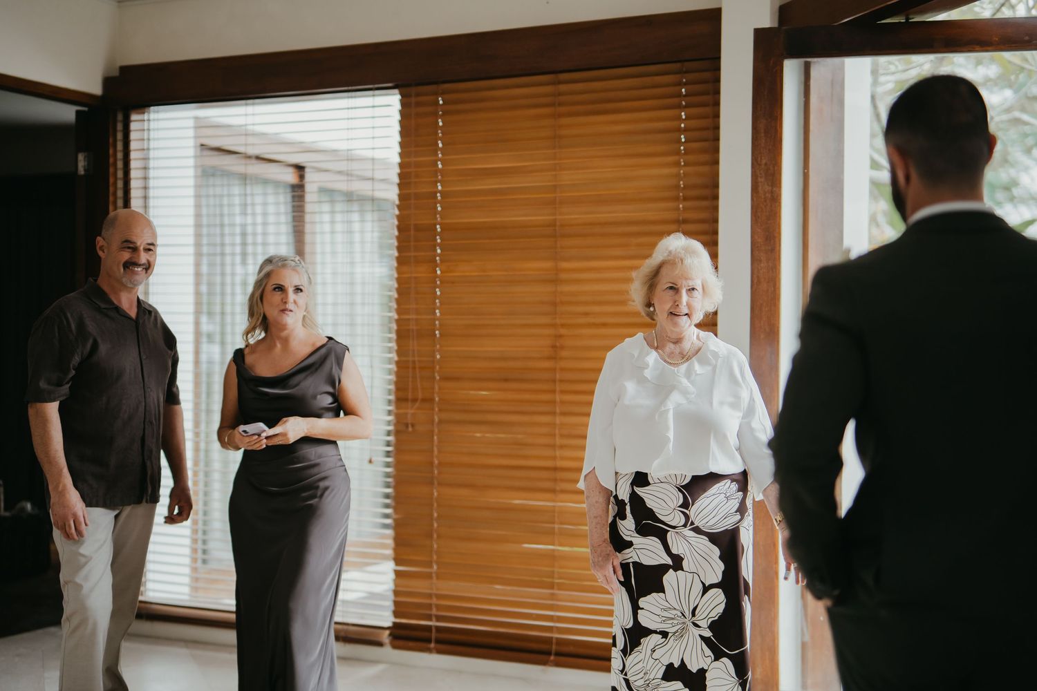 Family members gather indoors to greet a groom before his wedding ceremony near wooden blinds.