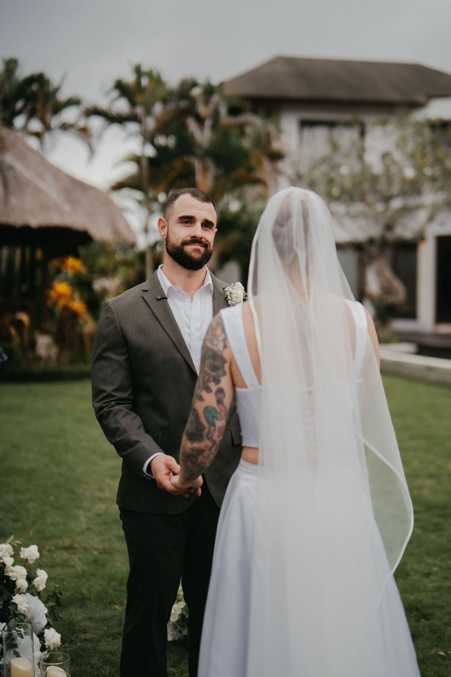 Groom with tattoos in dark suit holds bride's hands during outdoor wedding ceremony in tropical garden.