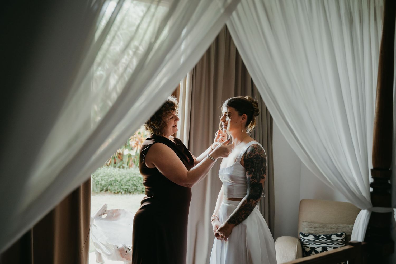Woman helps tattooed bride with makeup near sheer curtained window in soft natural light before wedding.