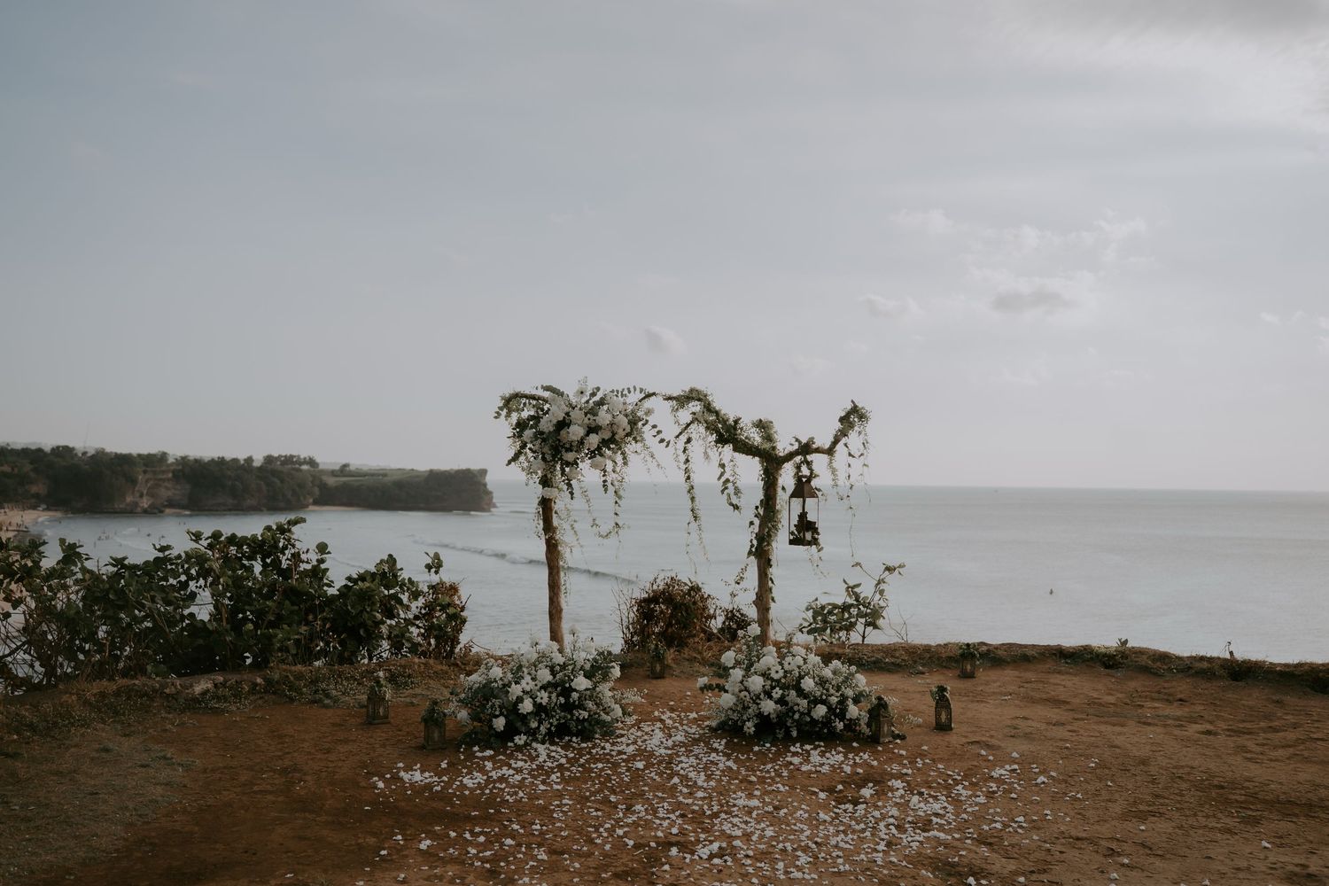 A rustic floral wedding arch stands on a coastal cliff overlooking the ocean under moody gray skies.