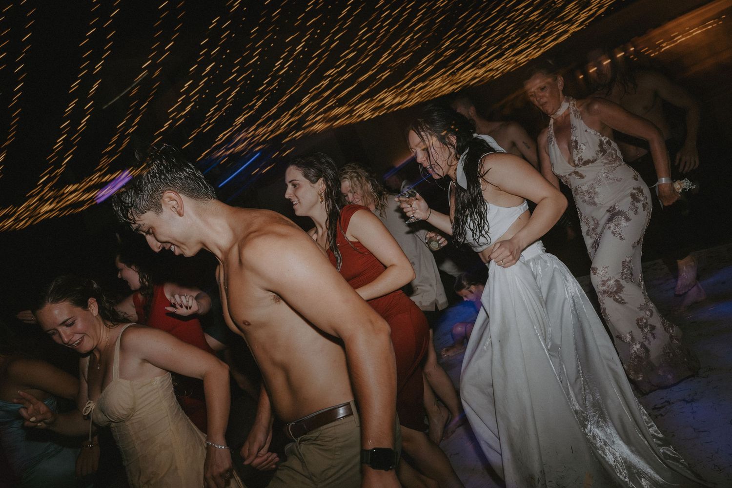 A group of people dancing in a line at a party with string lights overhead.