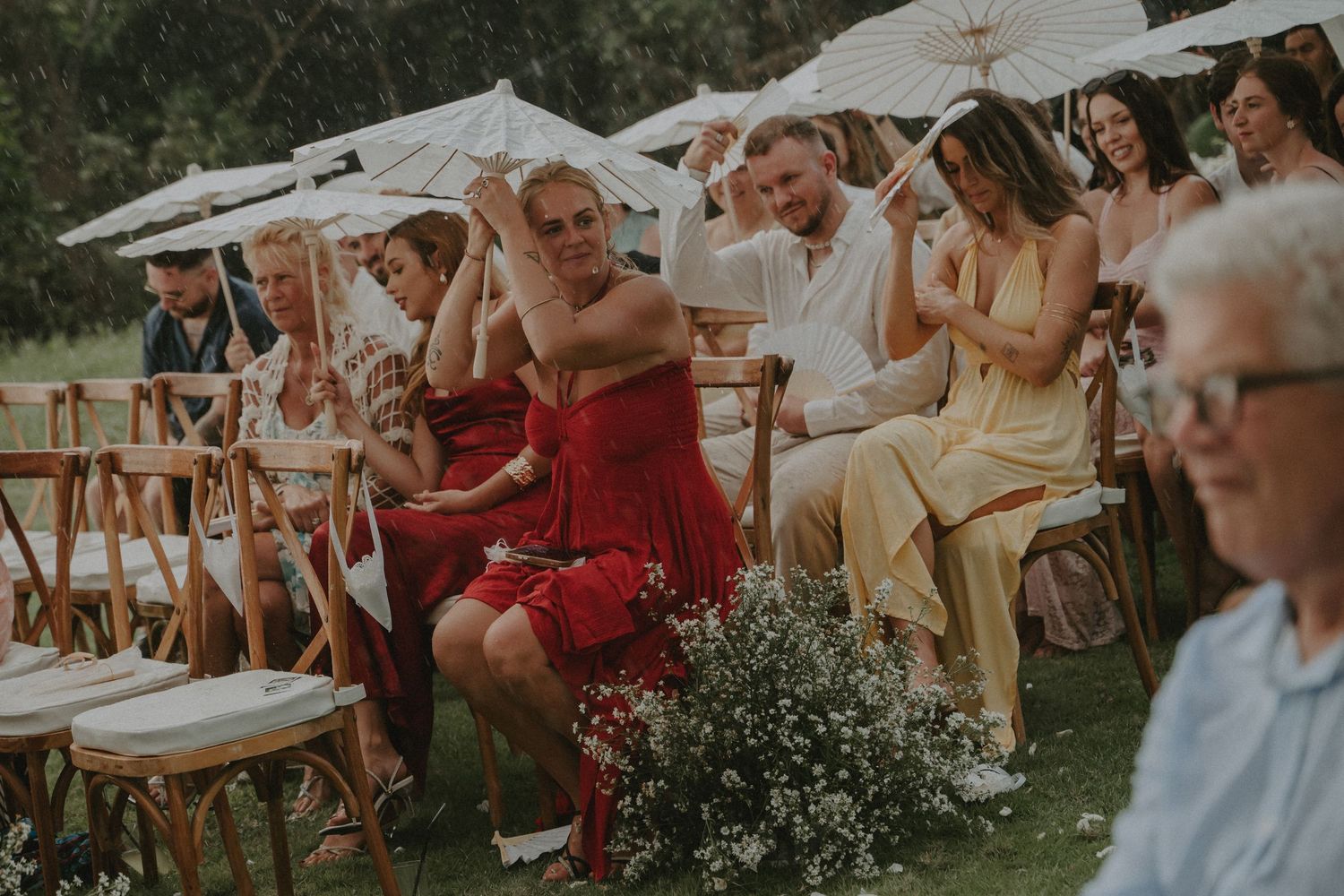 Wedding guests sit outdoors on white chairs during a summer garden ceremony with umbrellas for shade.