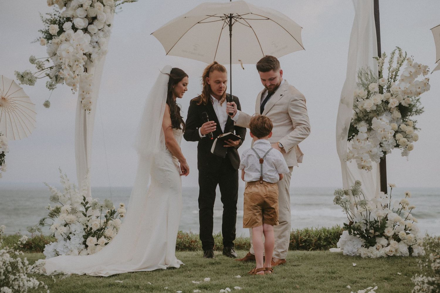 A romantic beachside wedding ceremony with white floral arches and guests gathered on a sandy shore.