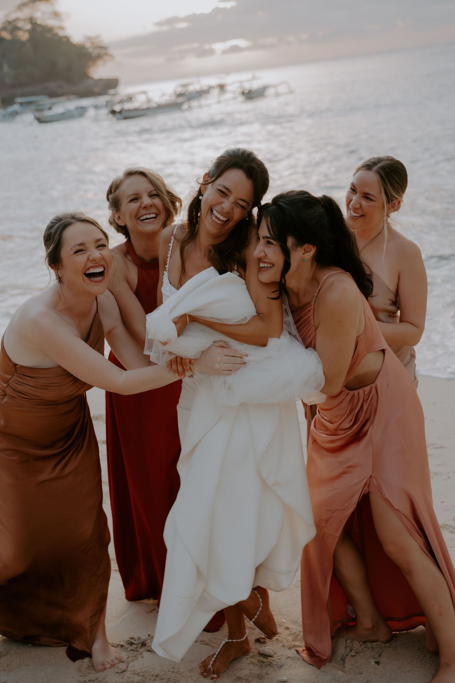 A group of bridesmaids in rust-colored dresses laugh together with the bride on a beach at sunset.