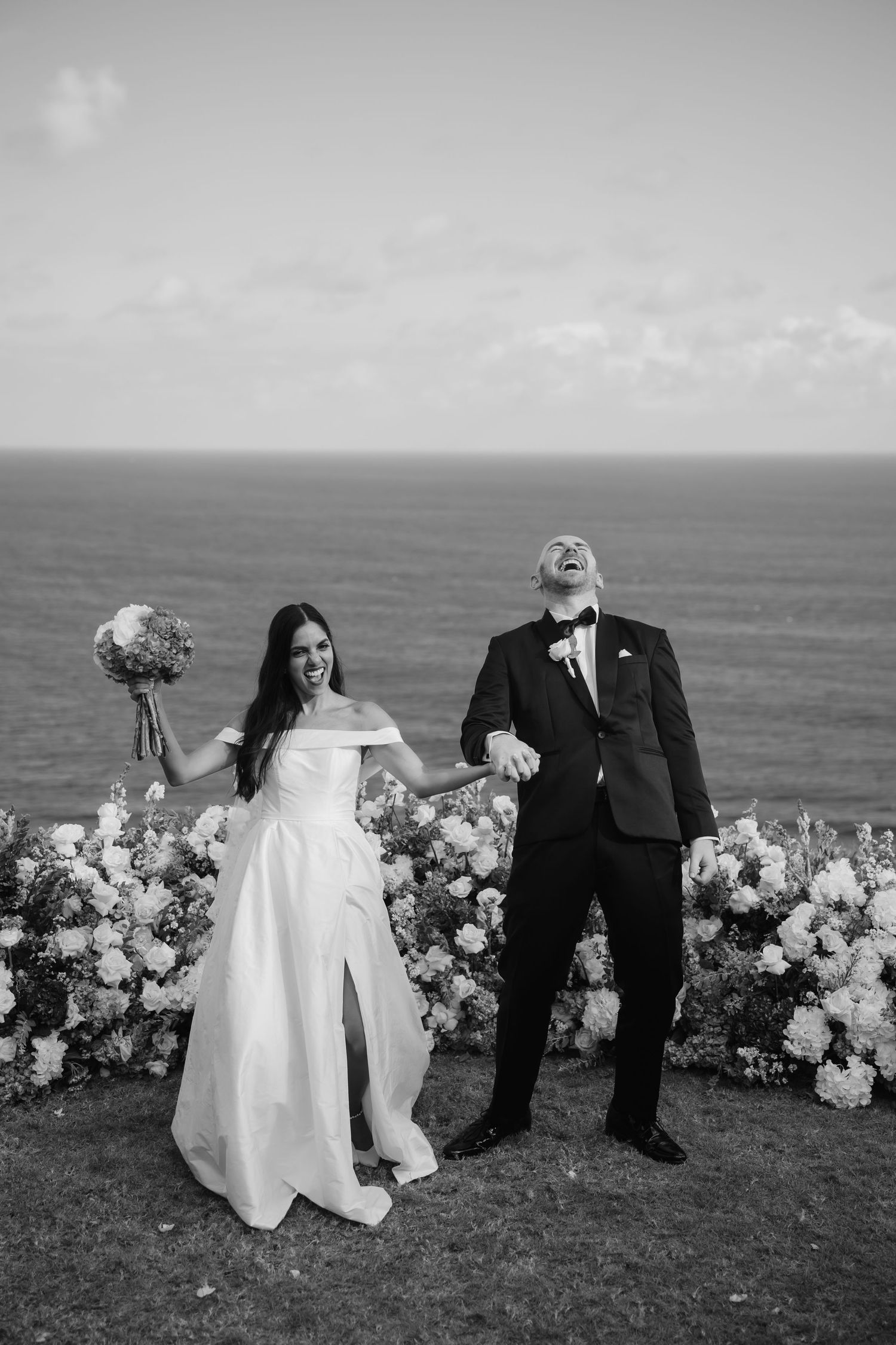 Wedding couple poses with floral arrangements on a dramatic oceanside cliff in black and white.