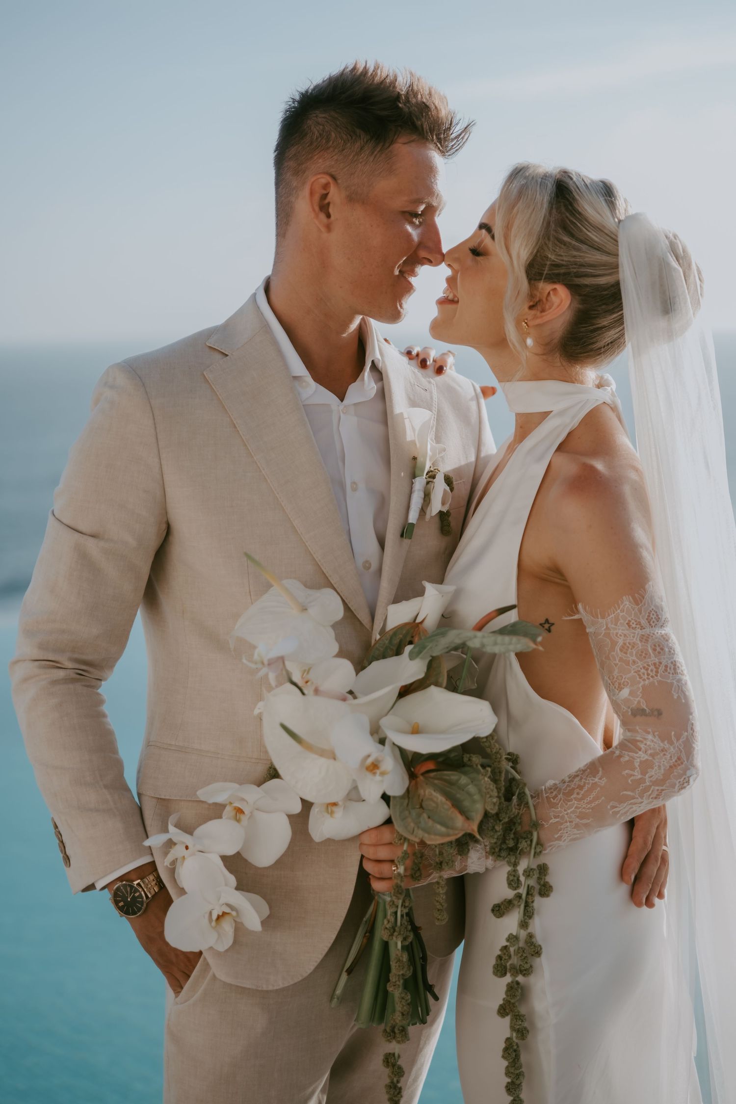Newlyweds share tender moment with white orchid bouquet against ocean view.