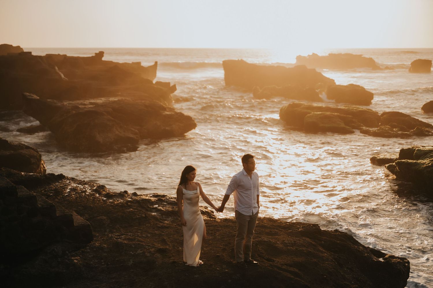 Couple holding hands on rocky coastal cliffs at golden sunset with ocean waves in background.