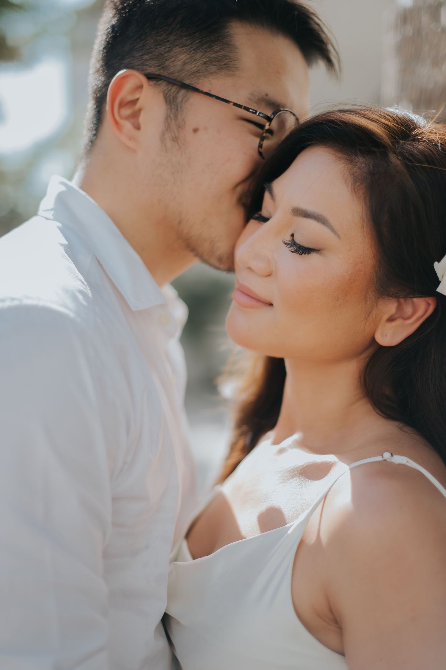 A couple in white attire shares a tender moment, the man gently kissing the woman's forehead in soft sunlight.