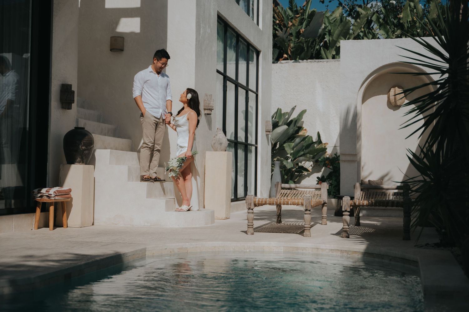 Couple standing on steps by a luxury pool in a white Mediterranean-style villa surrounded by tropical plants.