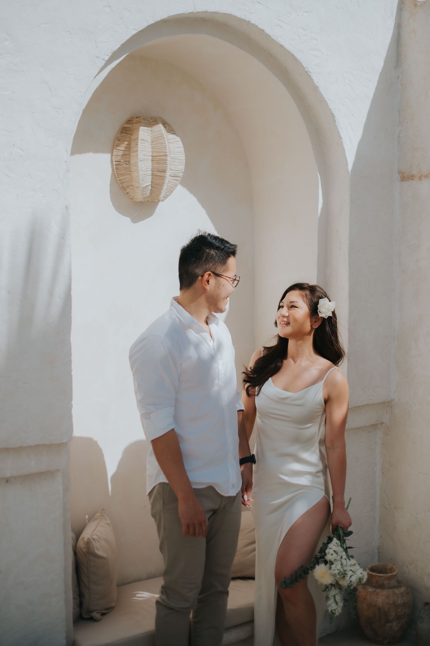 Couple in white attire posing under an arch in a Mediterranean-style setting for a romantic pre-wedding shoot.