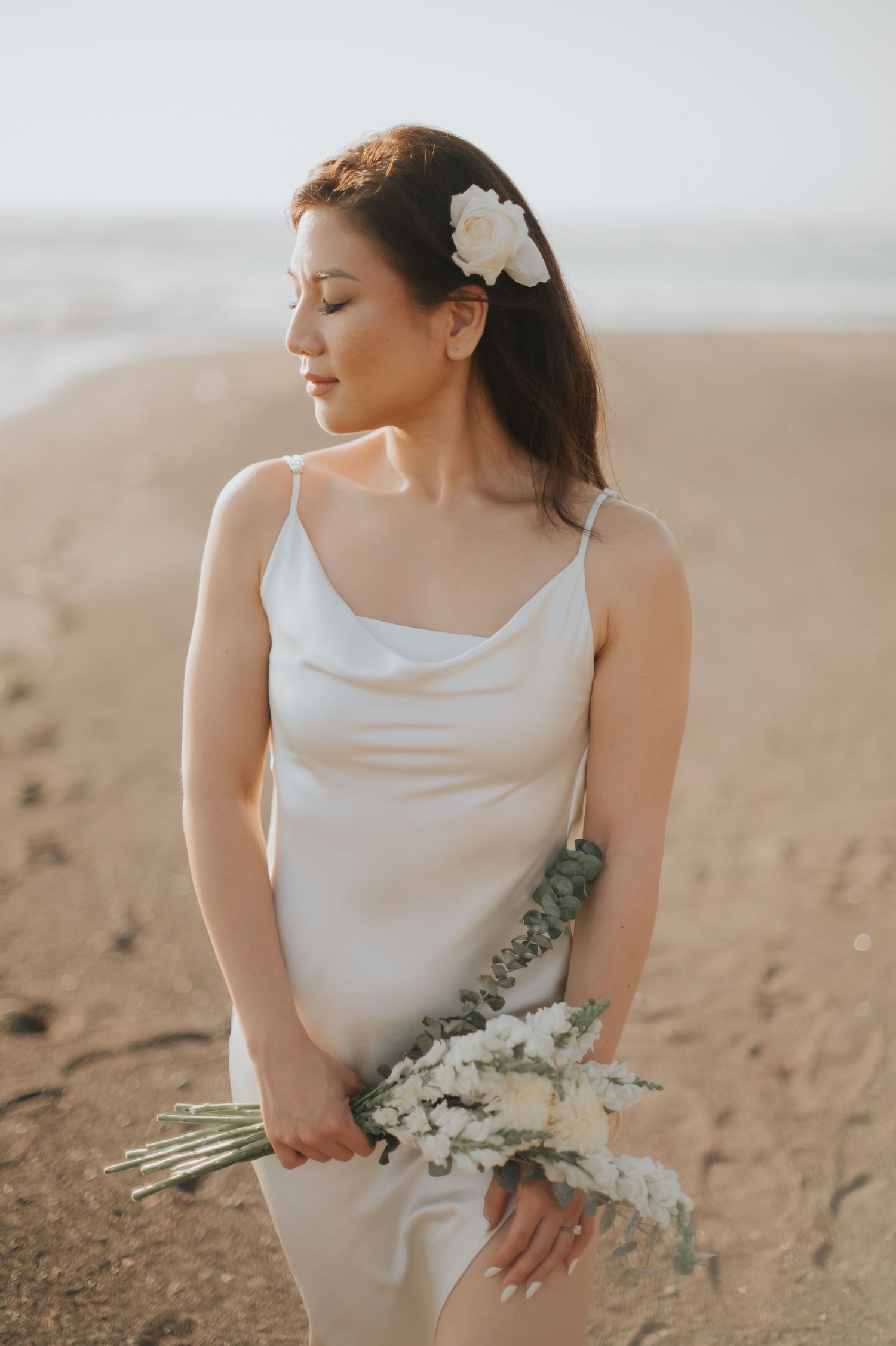 Bride in white satin slip dress holding floral bouquet on sandy beach, white flower hair accessory, serene expression.
