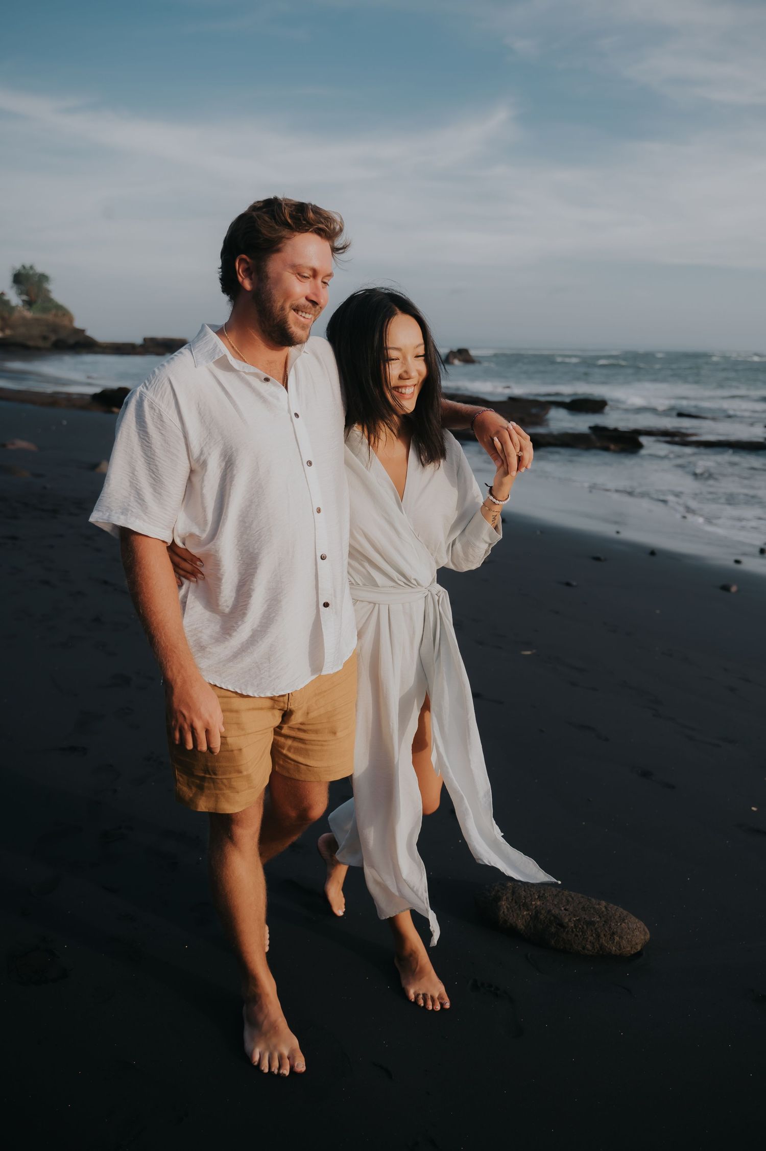 A couple stands barefoot on a black sand beach at sunset, dressed in casual white and beige clothing.