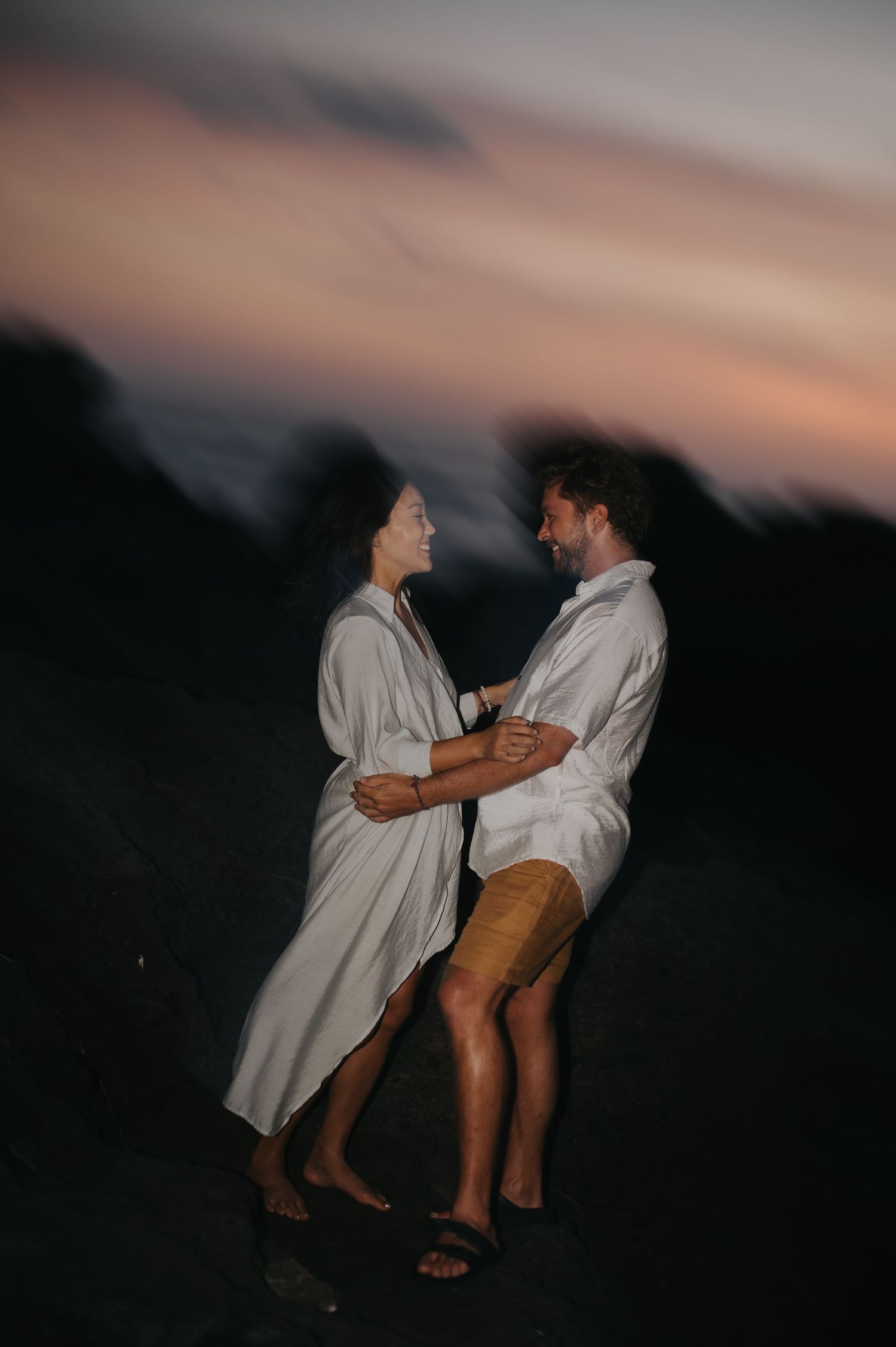 A couple in white shirts and shorts dances together at sunset creating silhouettes against the darkening sky.
