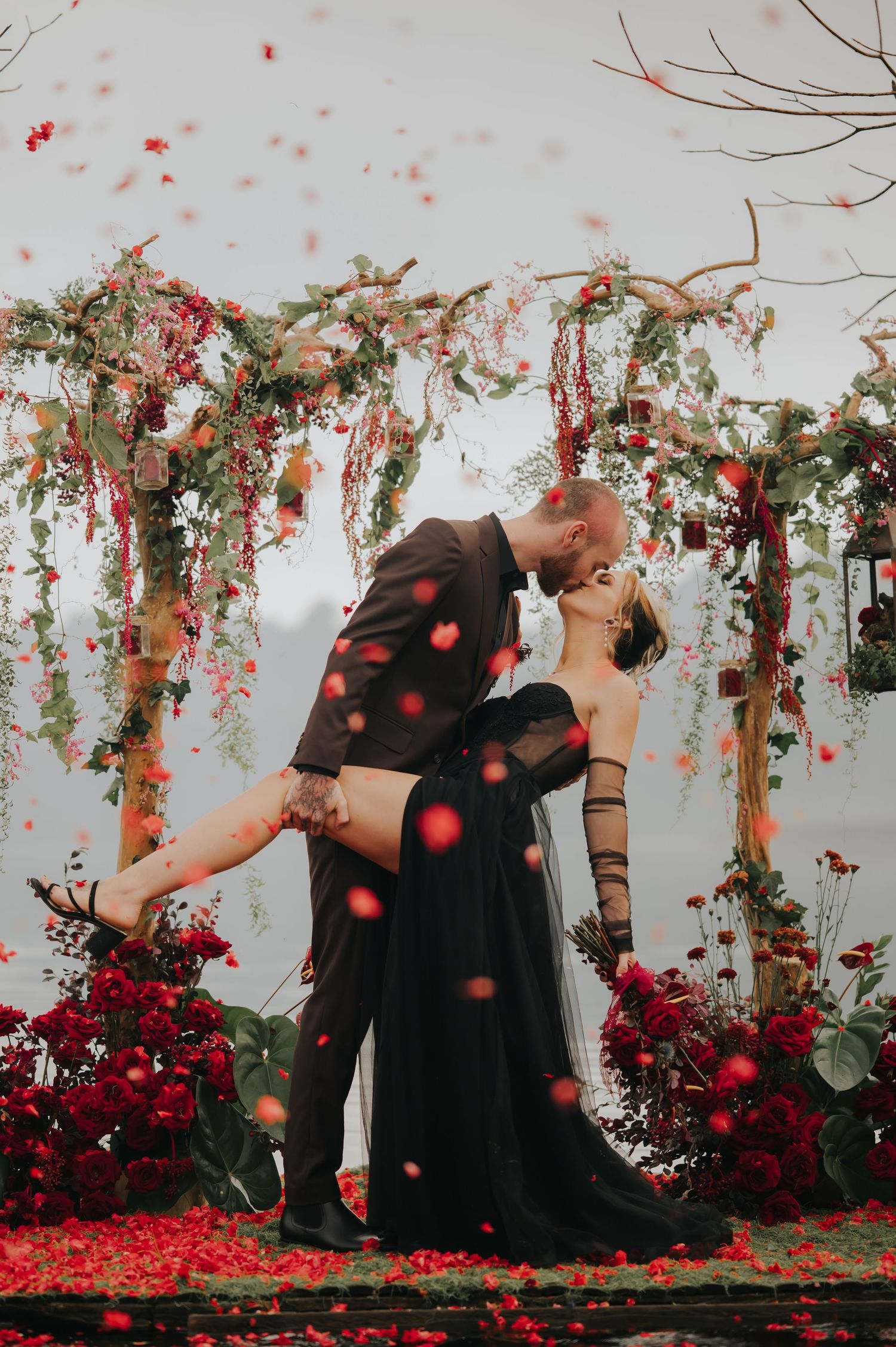Red rose petals fall around a couple embracing under a floral arch with white backdrop.