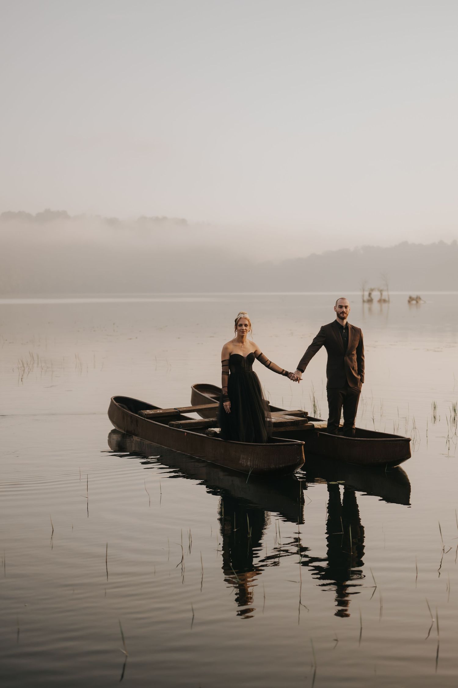 A couple sits in wooden canoes on a misty lake at dawn with reflections on the calm water.