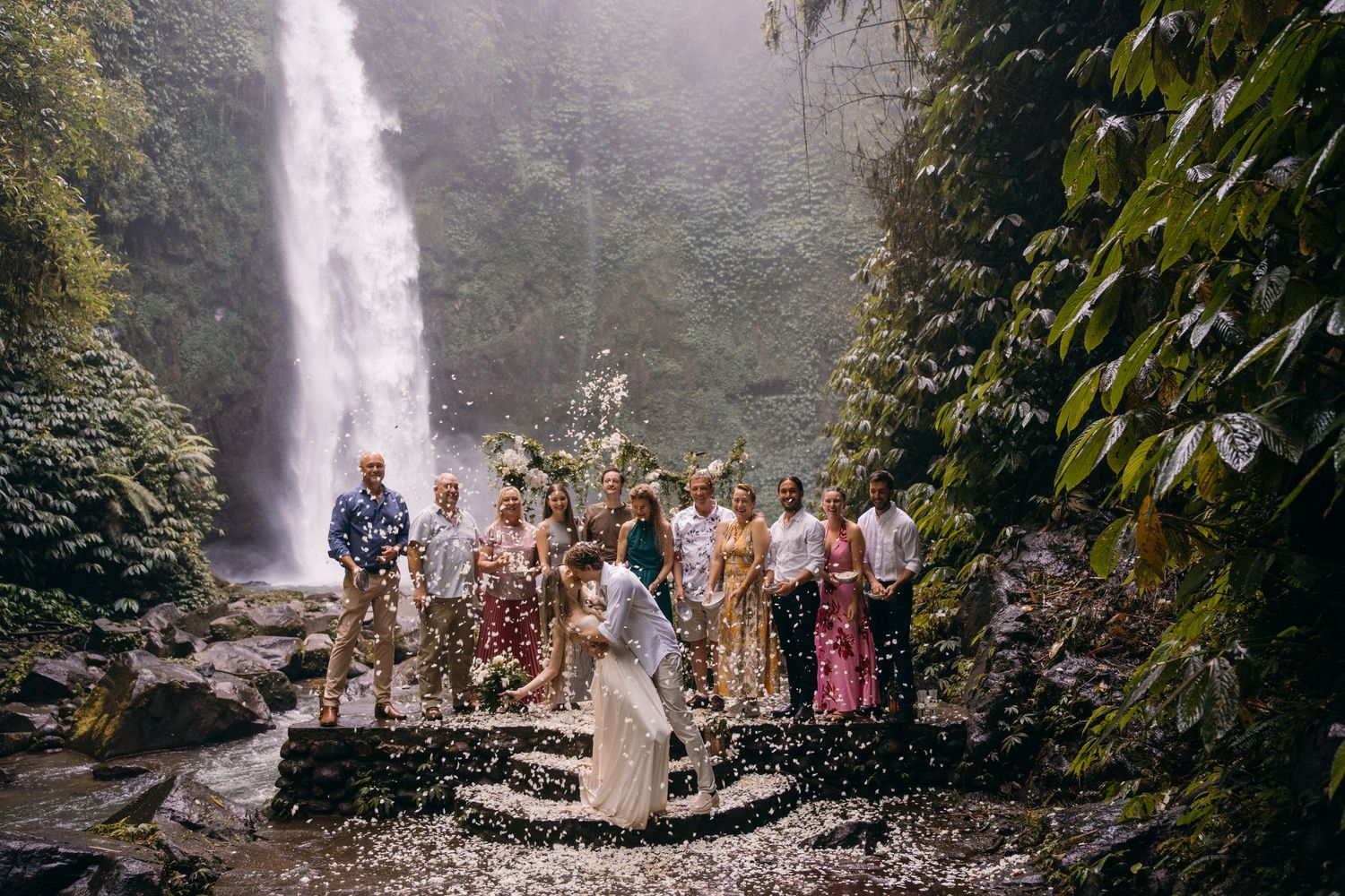 Wedding party poses in front of majestic waterfall in tropical forest setting during outdoor celebration.