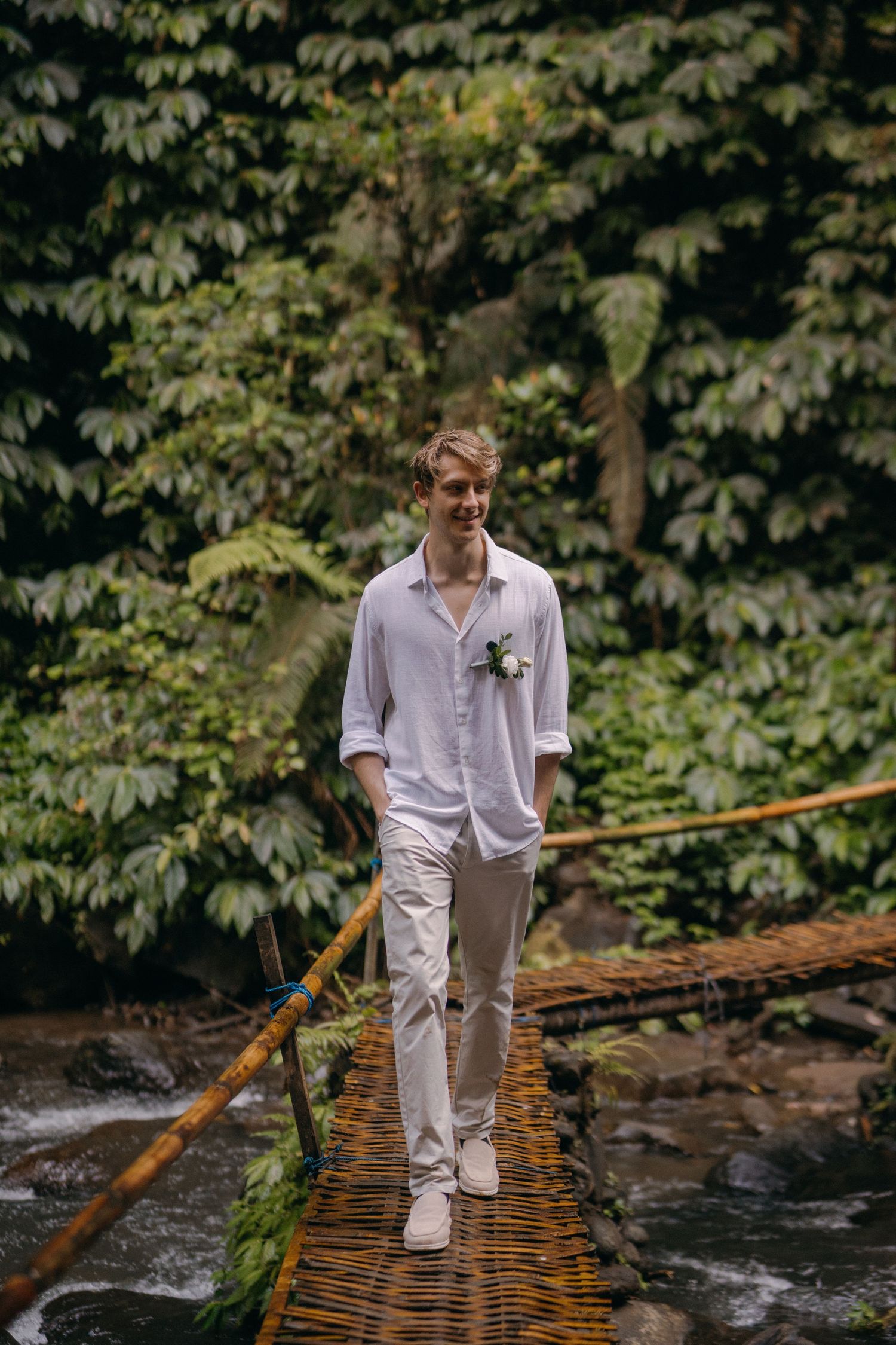 A person in white clothing carefully crosses a rustic wooden bridge over a flowing stream in a lush forest setting.