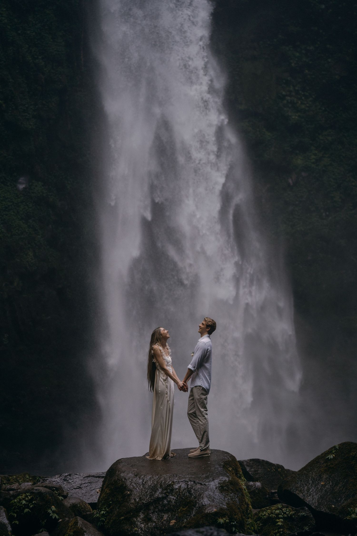 A couple stands silhouetted against a majestic waterfall backdrop in a dramatic engagement photo shoot.