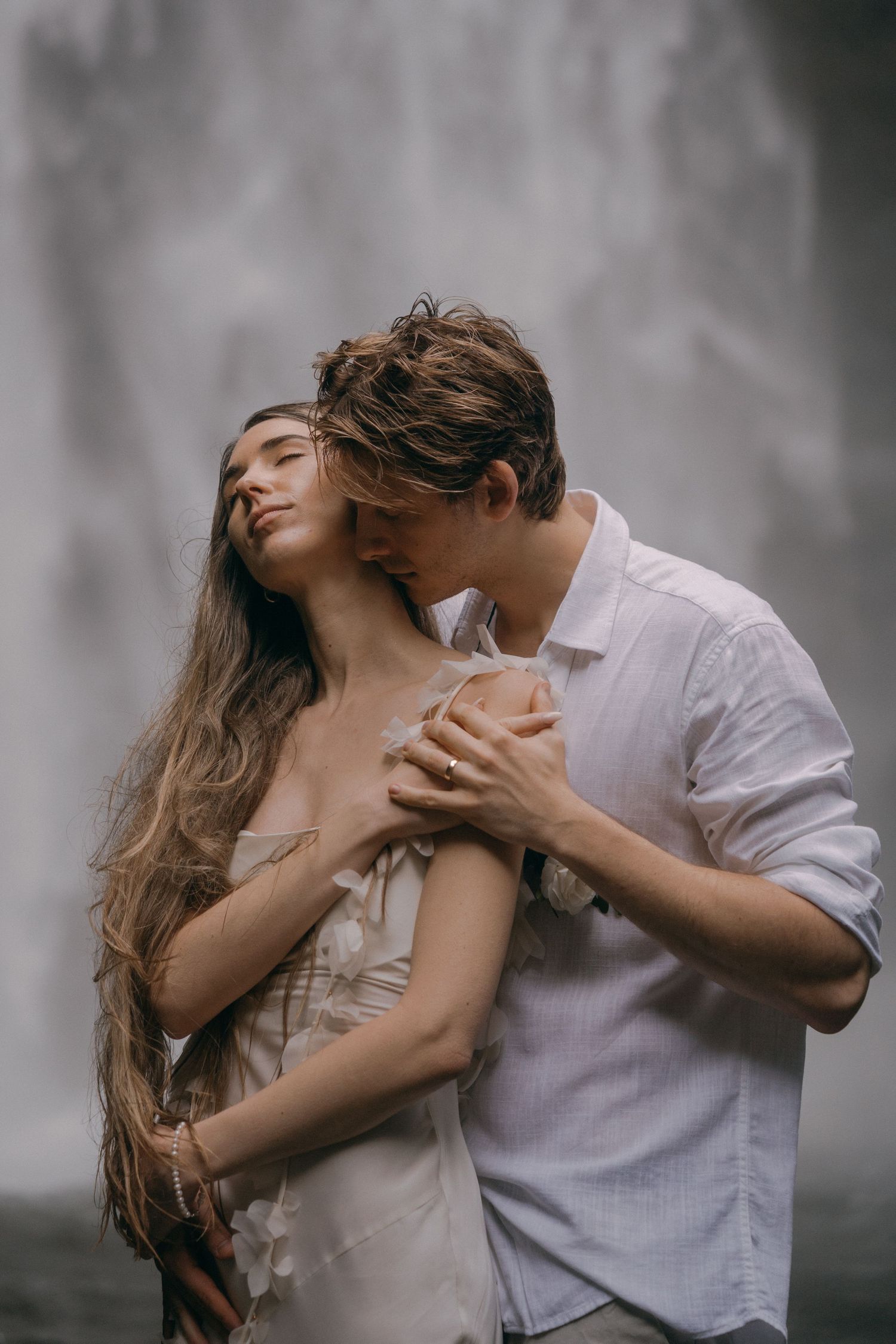 Romantic couple embraces passionately in front of misty waterfall backdrop with moody gray atmosphere.