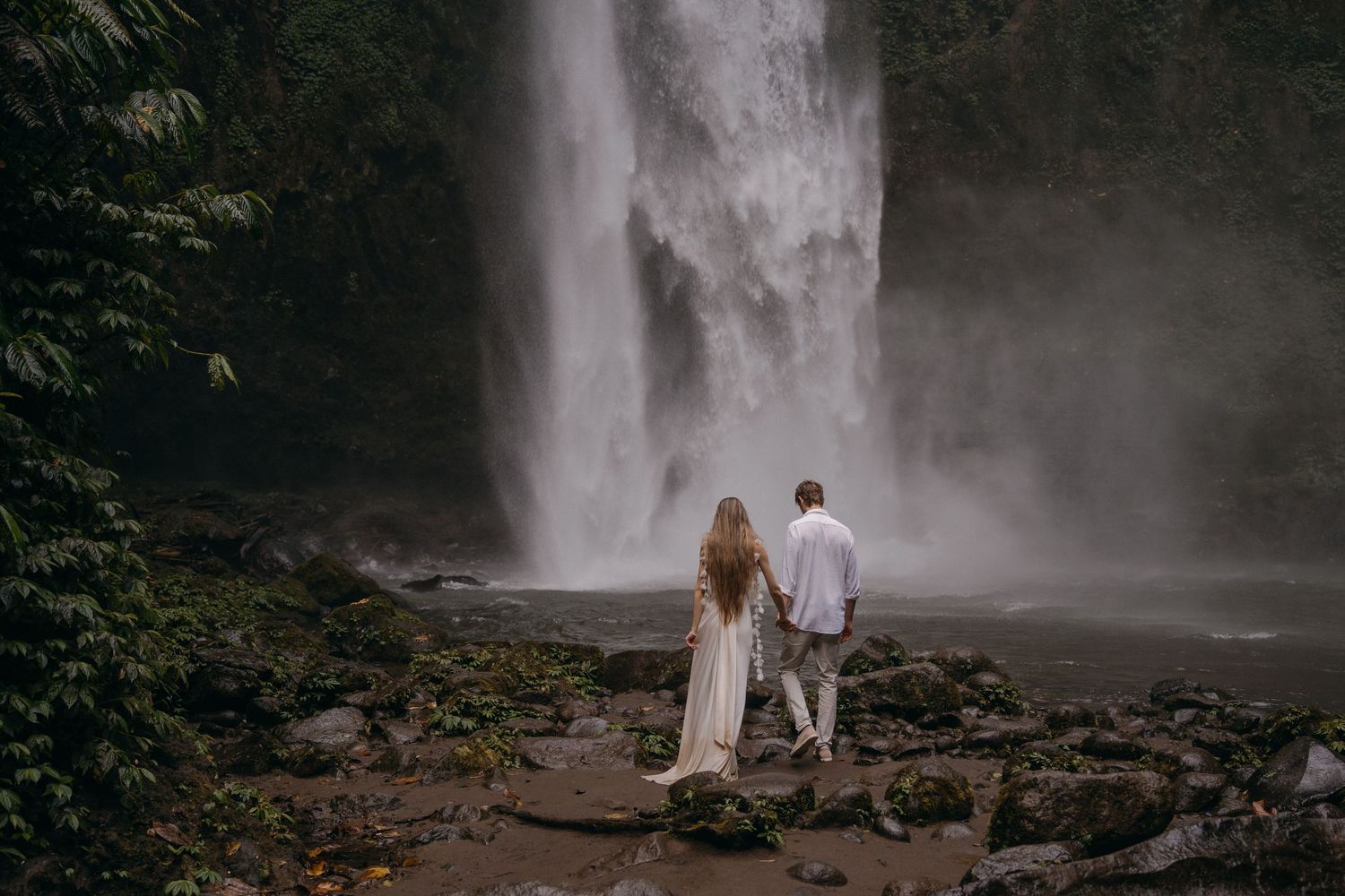 A couple stands near a misty waterfall surrounded by rocks and lush greenery in a moody natural setting.