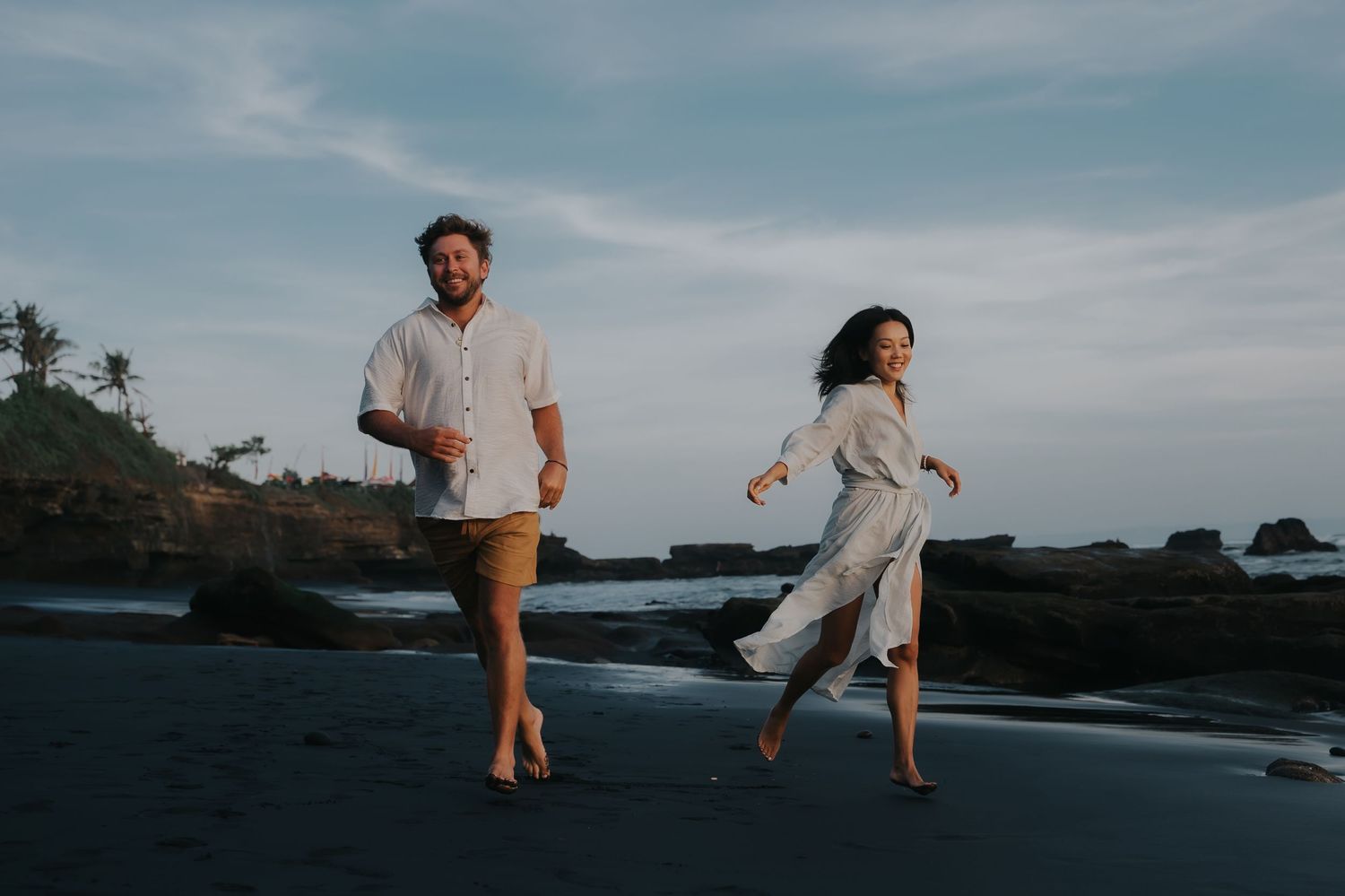 Couple walking along a rocky beach at sunset with waves crashing against the shore in the background.