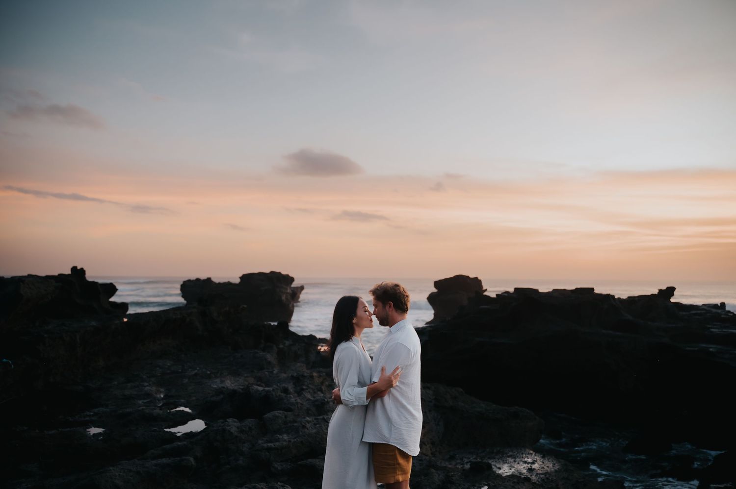 Silhouettes of people embracing on a rocky beach at sunset with dramatic orange and pink sky in the background.