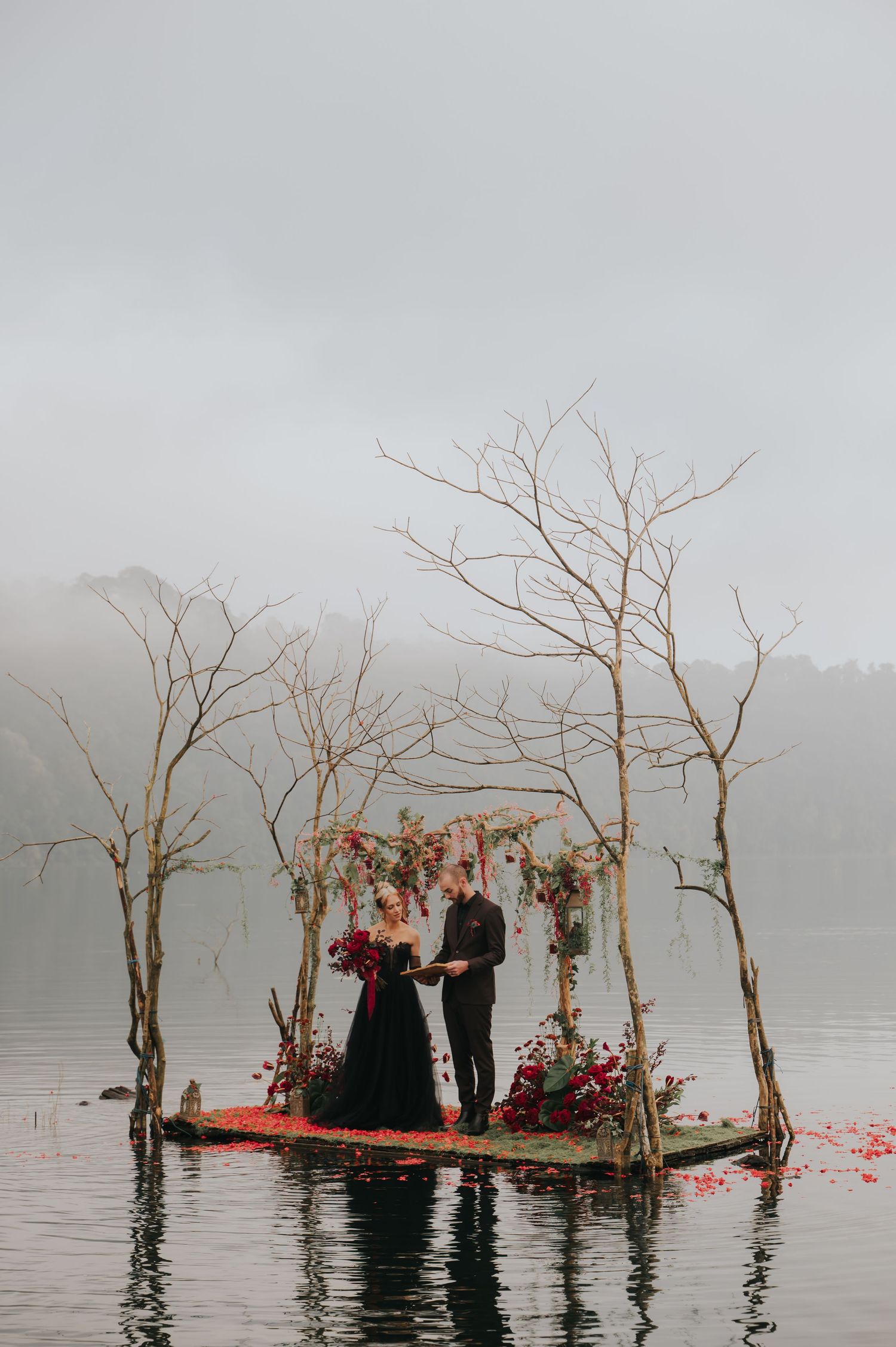 A moody wedding scene on a misty lake with bare trees and scattered red flower petals.