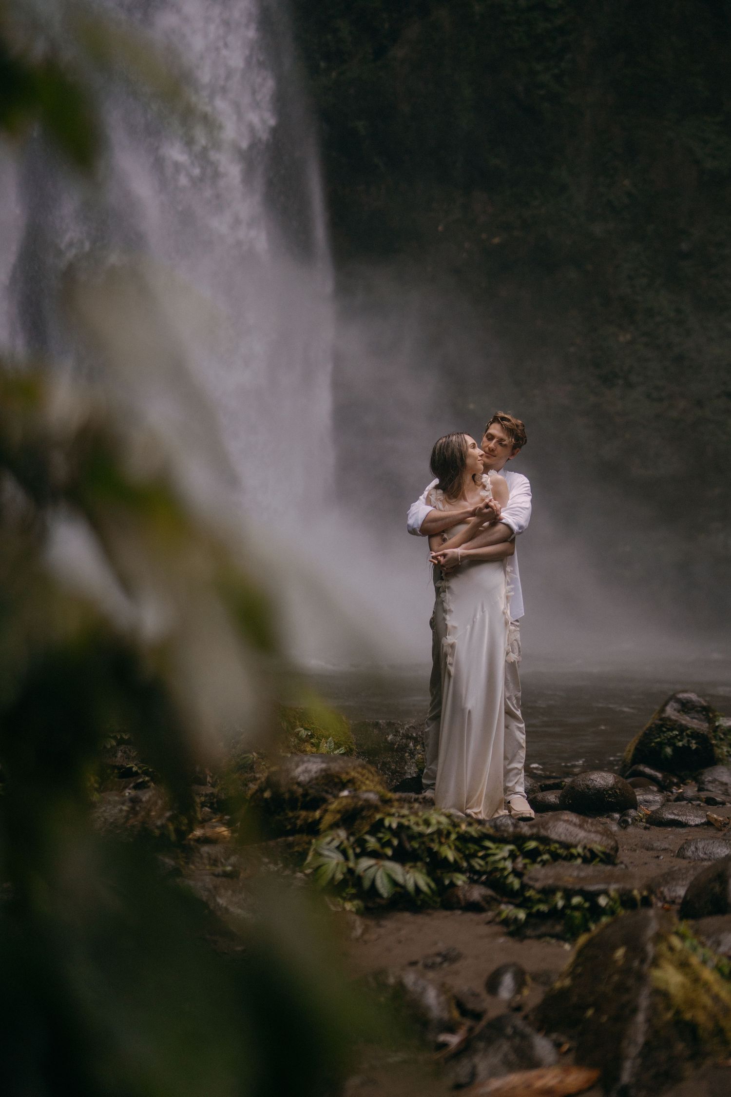 A figure in a flowing white dress poses dramatically against a misty waterfall backdrop in a moody natural setting.