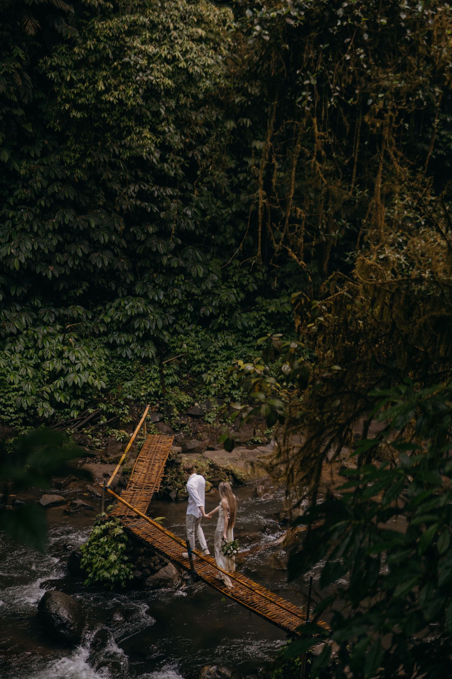 A couple walks across a wooden footbridge over a rocky stream in a lush forest setting during golden hour.