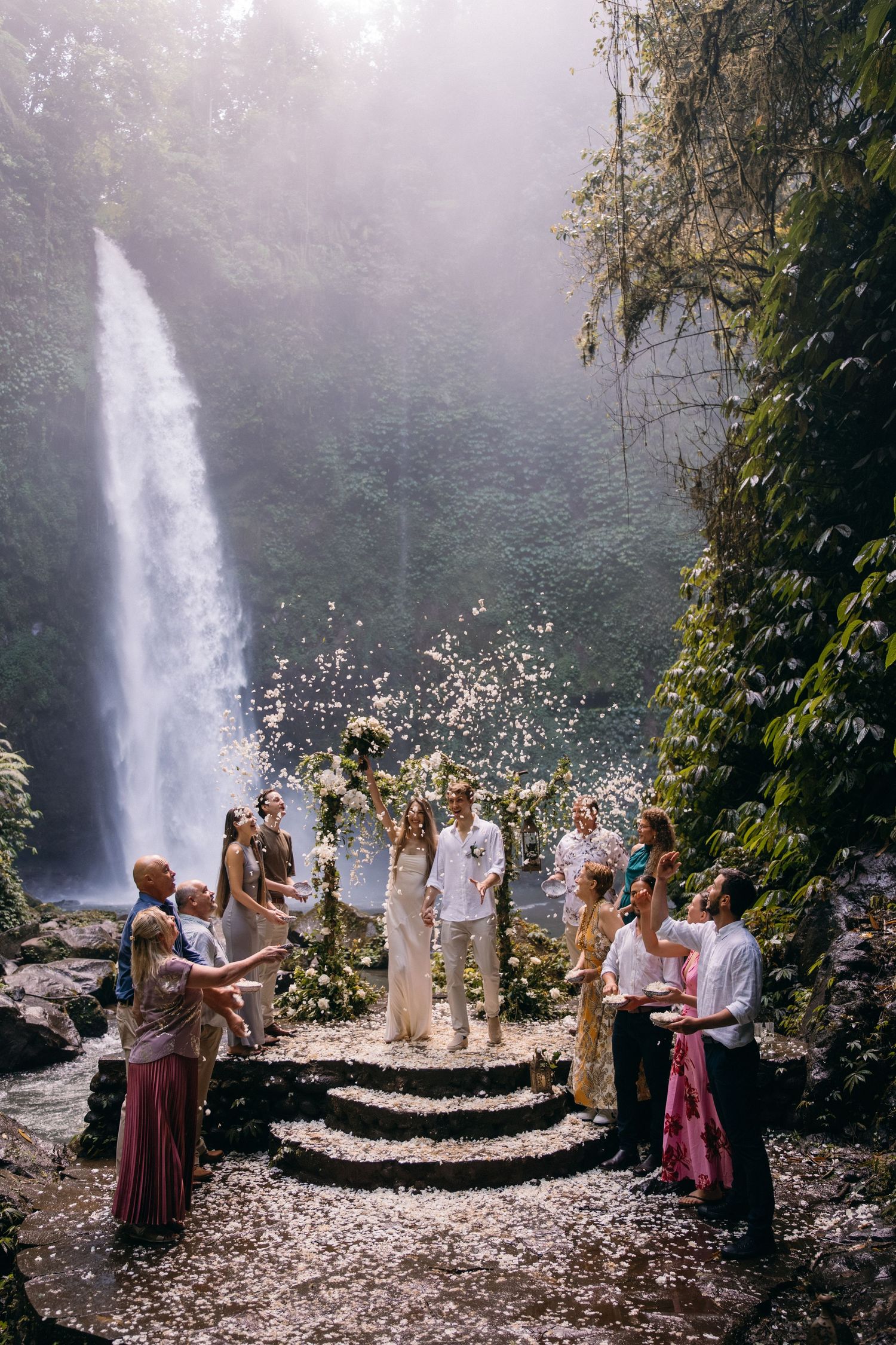A wedding ceremony takes place on stone steps in front of a majestic waterfall surrounded by lush tropical foliage.
