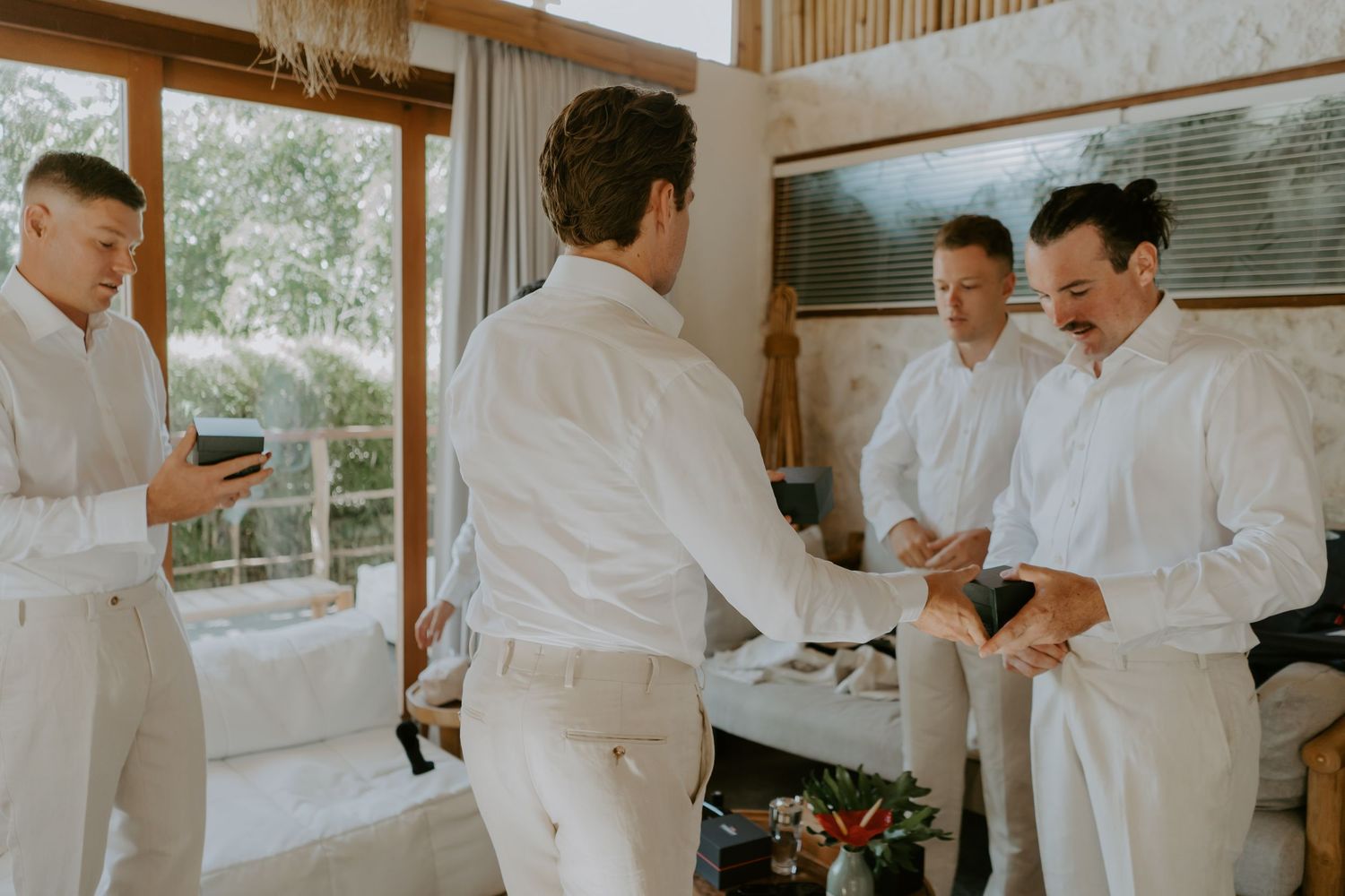 Groomsmen in matching white linen suits exchanging gifts and laughing together in a bright villa room.