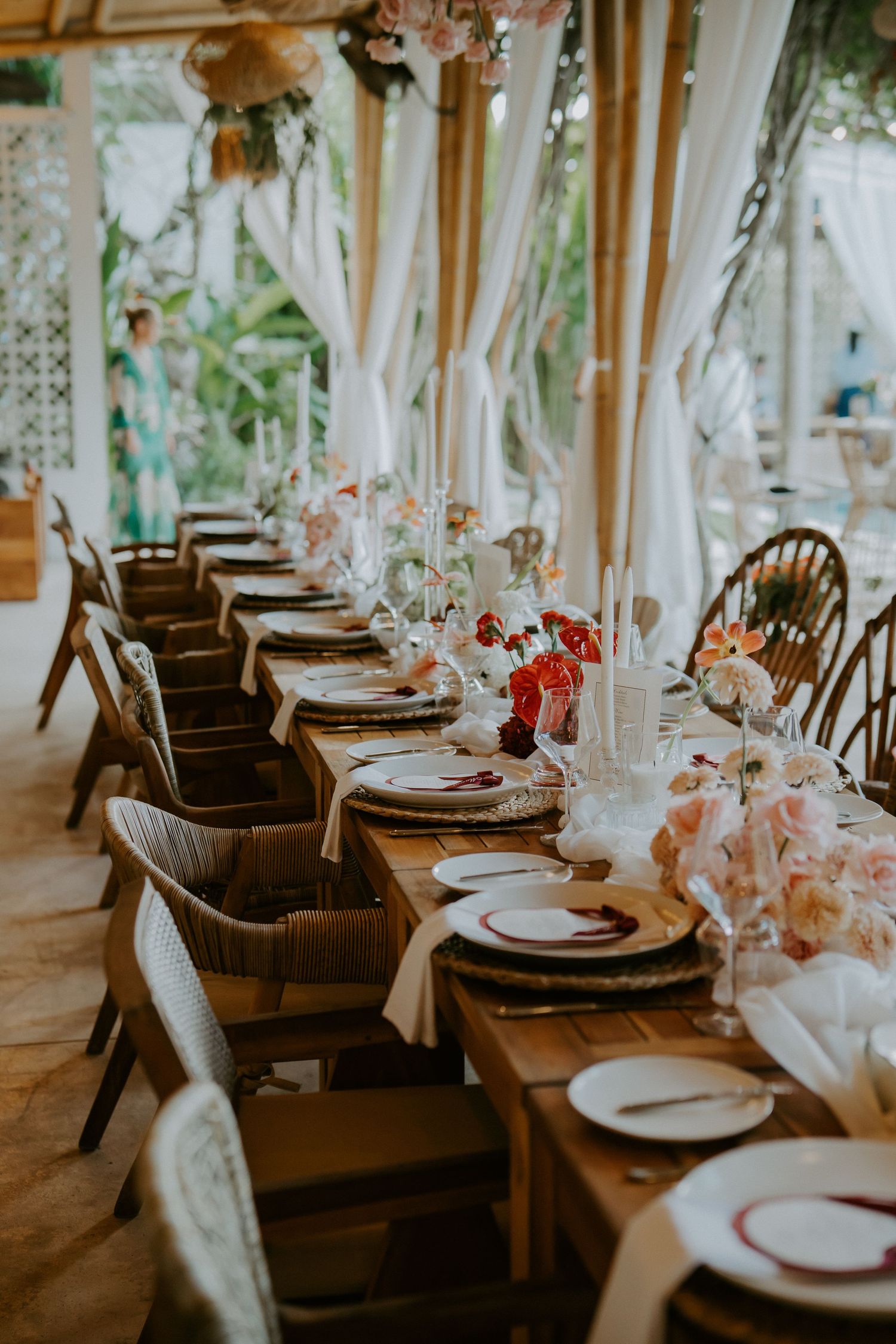 Long elegant wedding reception table with colorful floral arrangements, white candles, and wicker chairs under draped bamboo pavilion.