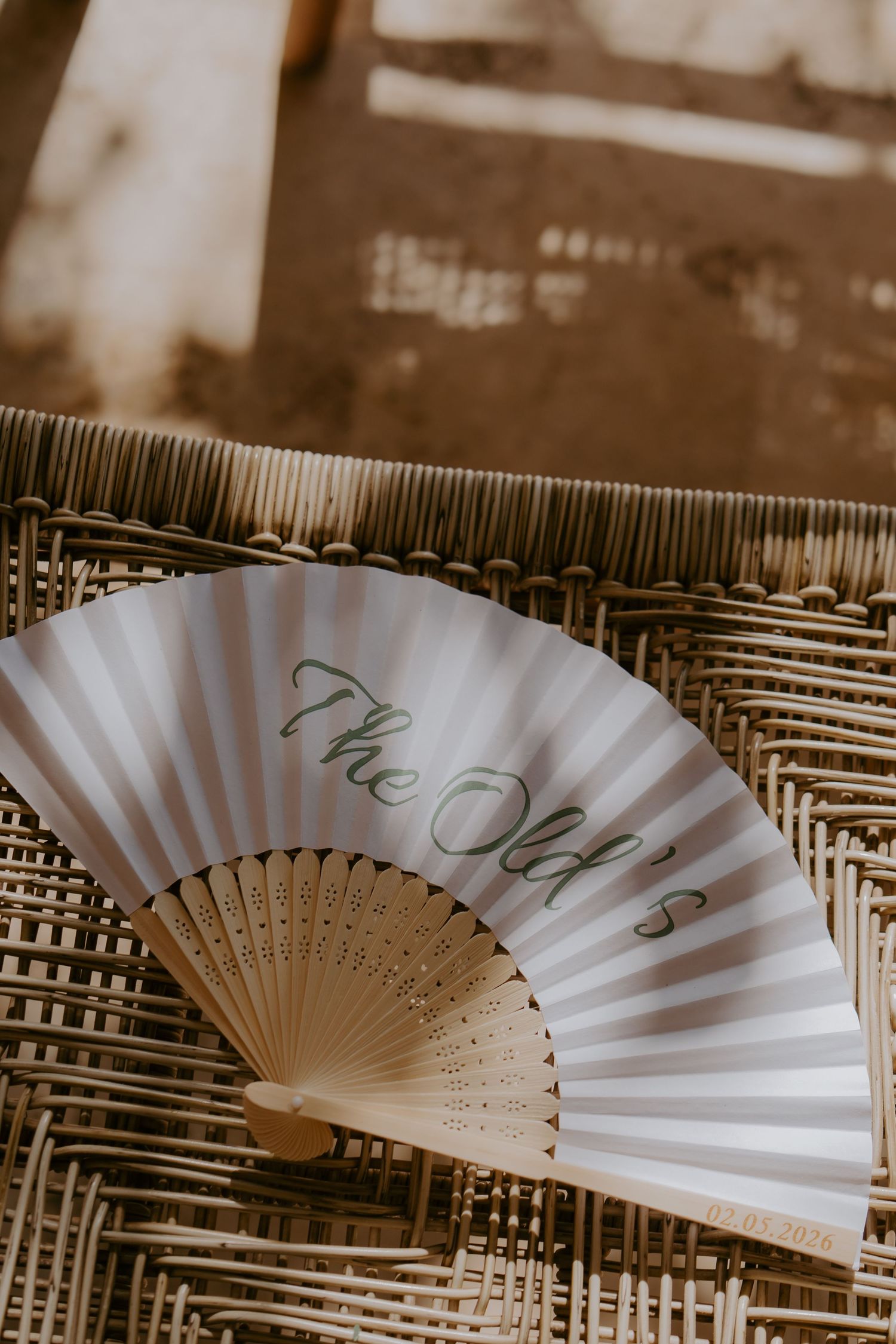 Elegant white hand fan with green cursive text reading The Olds resting on a woven rattan surface.