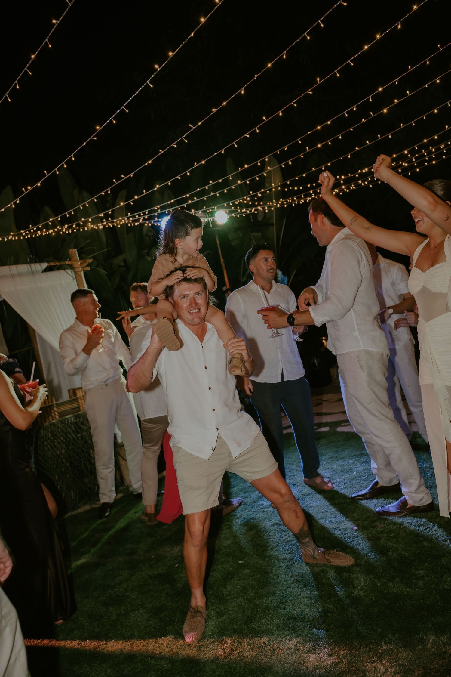 Energetic wedding guest dances with child on shoulders at lively outdoor nighttime wedding reception.