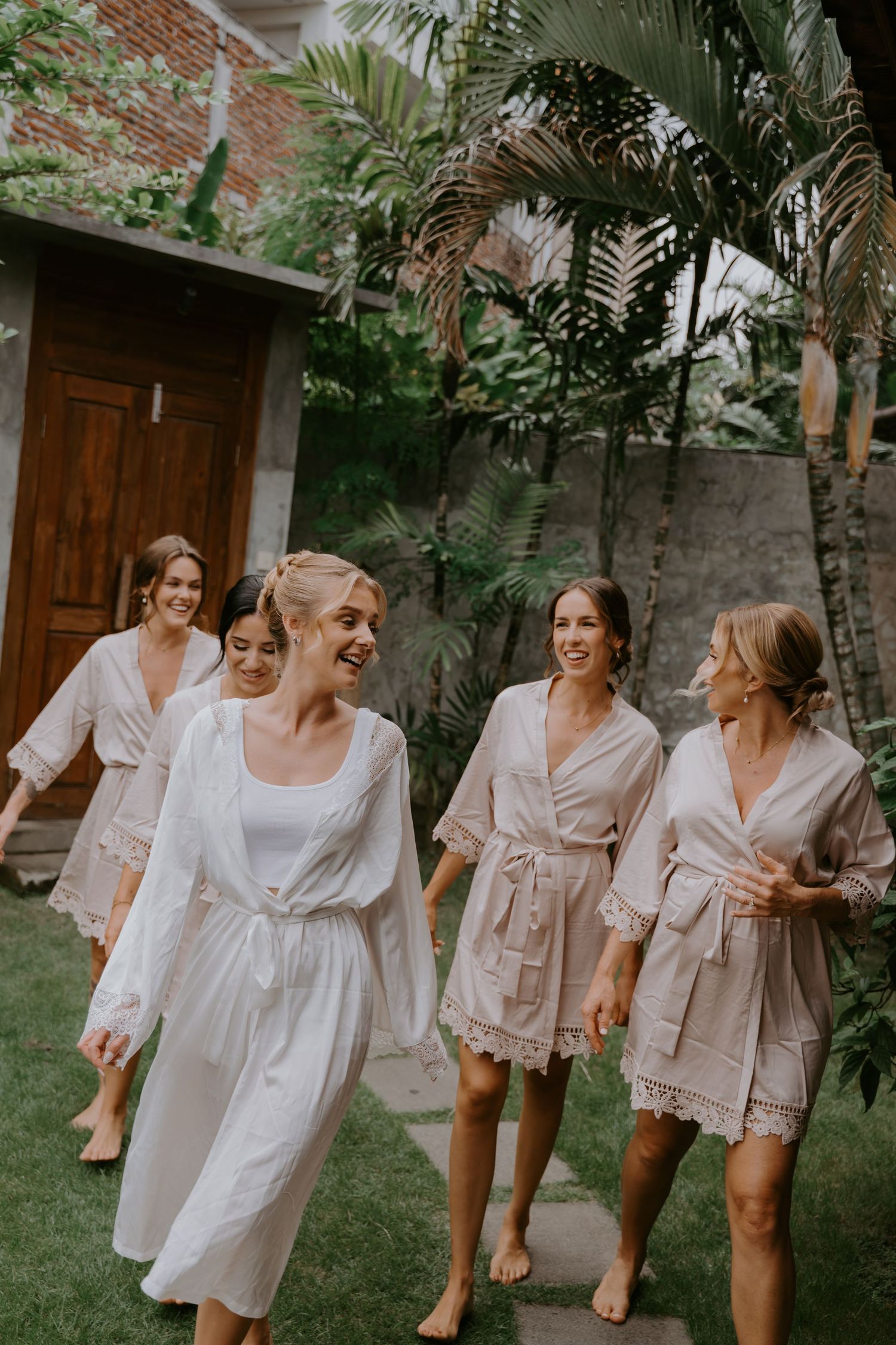 Bride in white robe leads bridesmaids in blush robes walking through tropical garden.