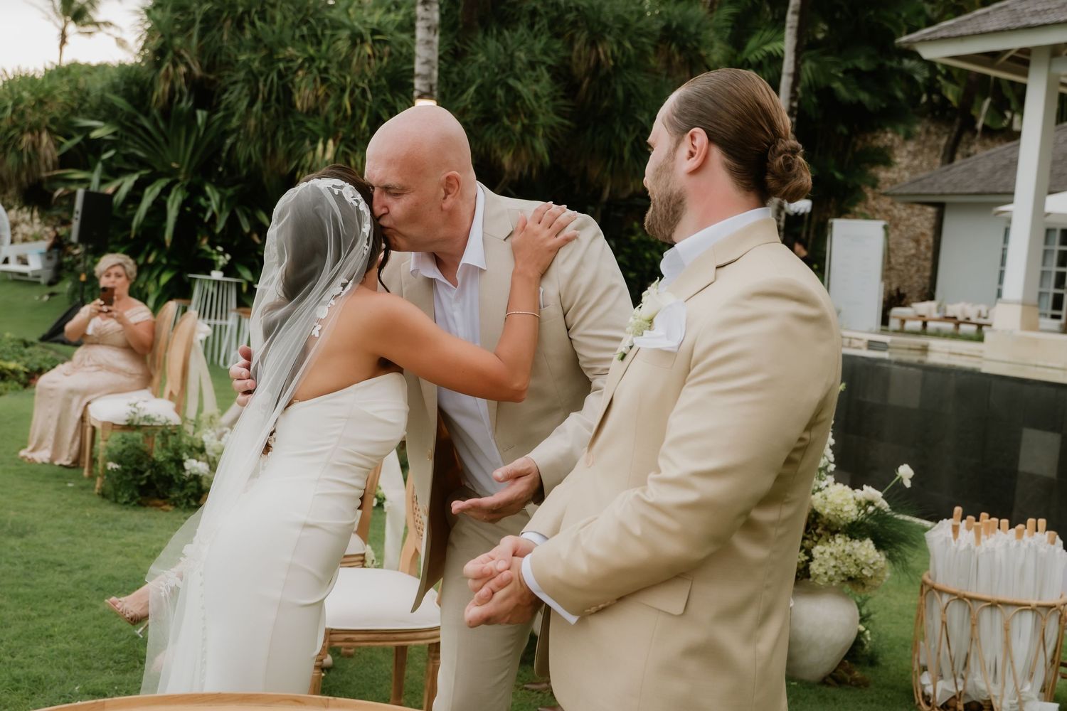 A bride shares a kiss with a guest while the groom stands nearby at the ceremony.
