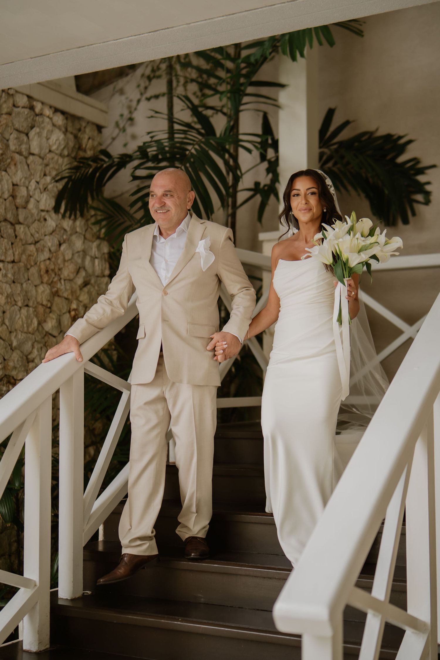 Bride in white gown holds bouquet of white lilies while descending stairs with man in beige suit.