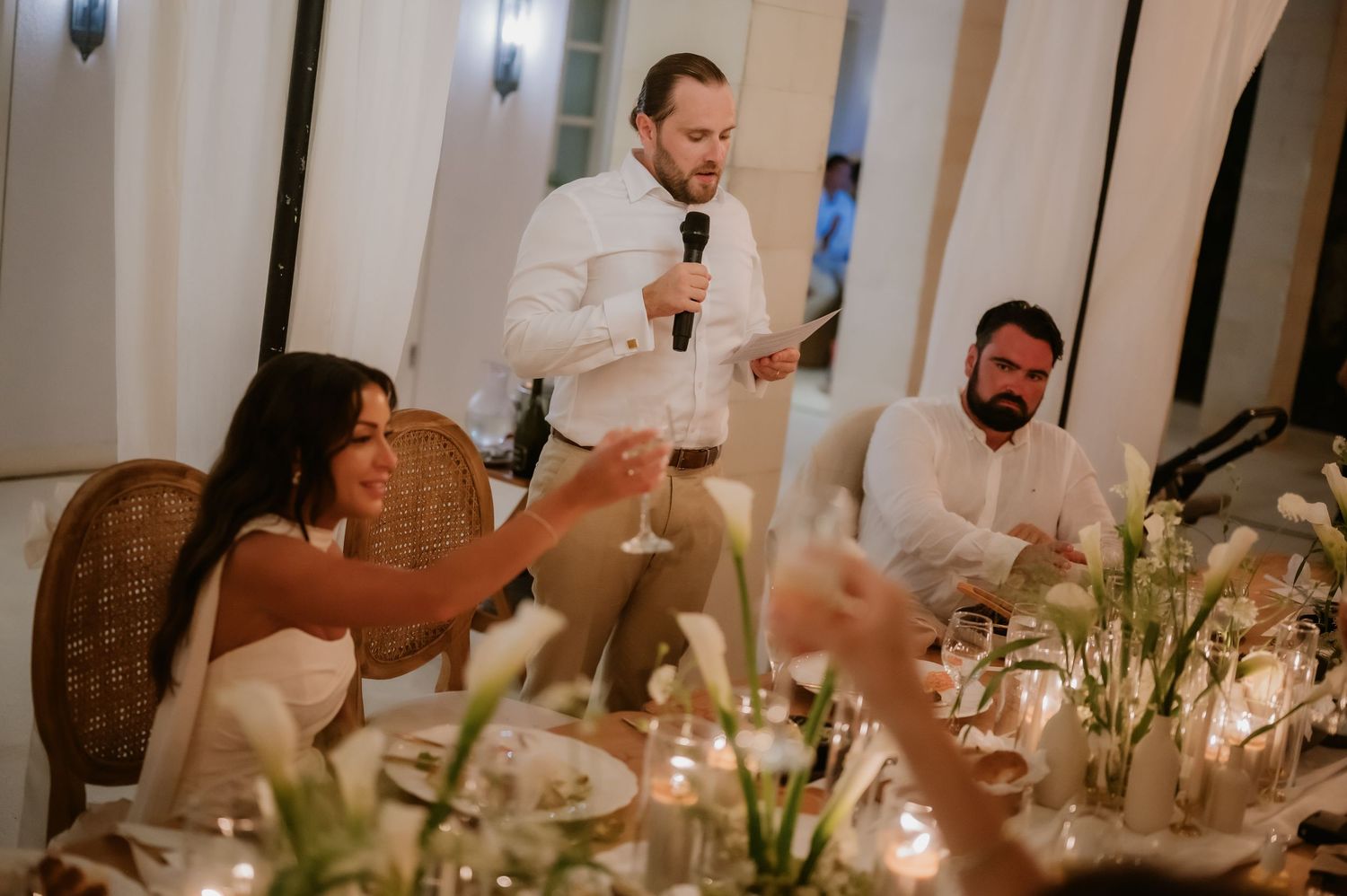 Groom gives a heartfelt toast with microphone as bride raises champagne glass at elegant wedding reception.