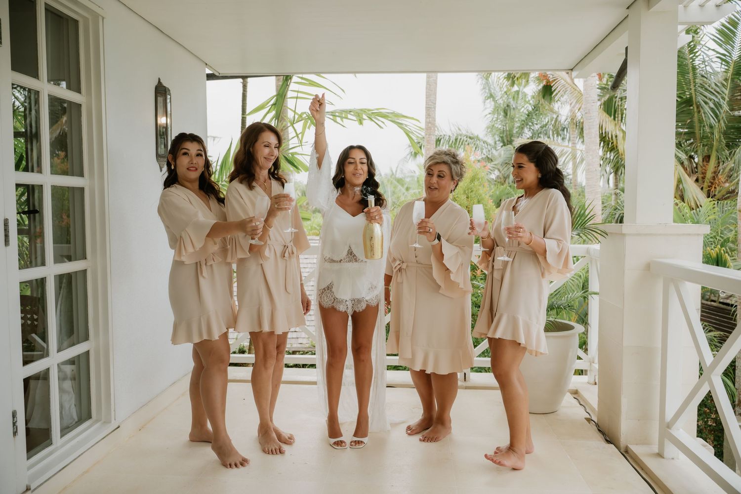 Five bridesmaids in matching beige satin robes laughing and holding champagne on a tropical villa porch.