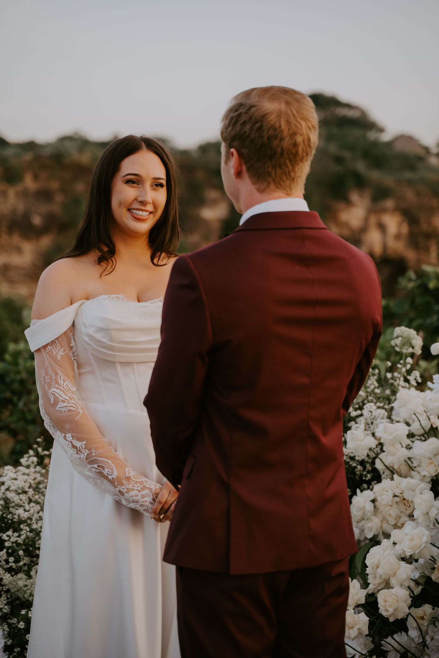 Radiant bride in lace wedding dress holds groom's hands during outdoor ceremony with white roses.
