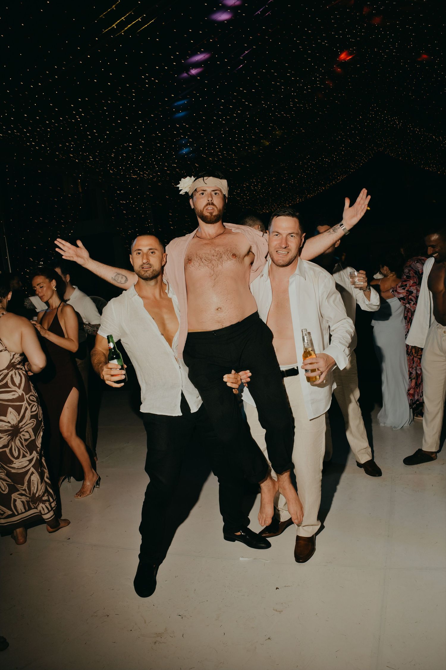 Three men celebrating on a dance floor at a wedding reception, two in white shirts and one shirtless with a floral crown.