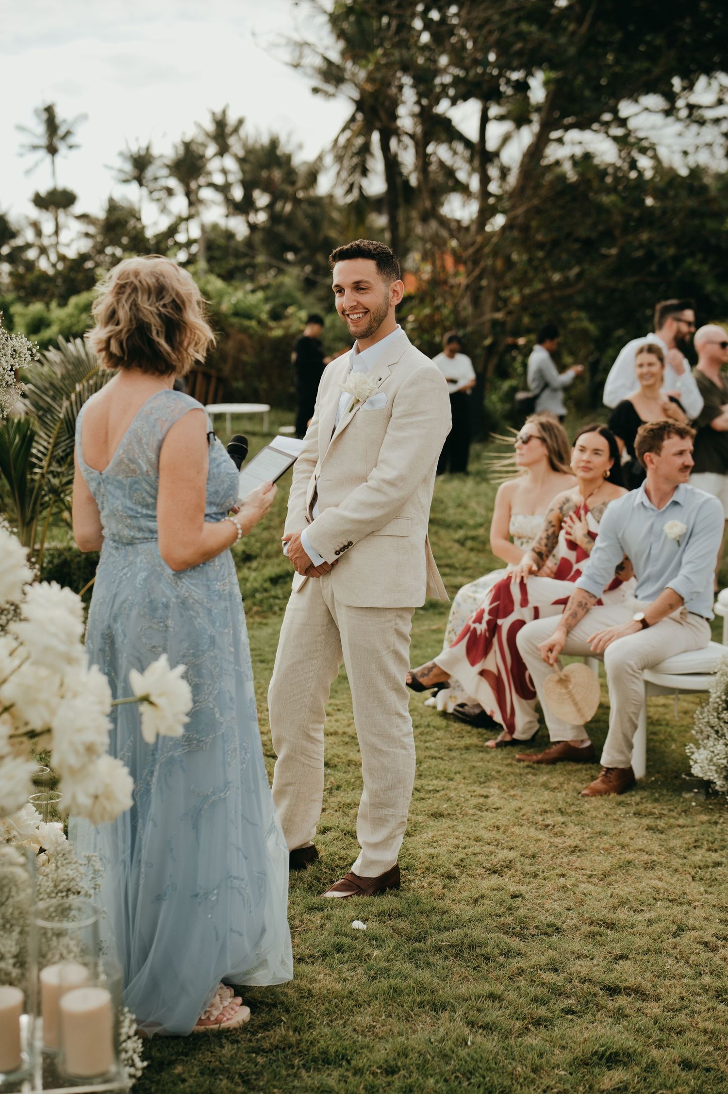 Groom in beige suit smiling at outdoor wedding ceremony with guests seated on lawn.