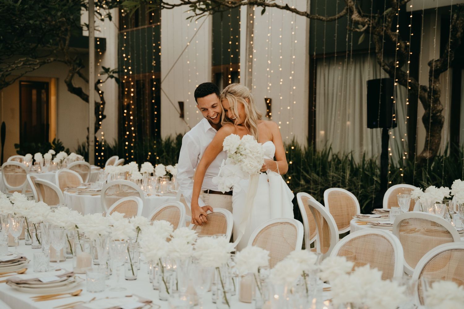 Newlyweds share a tender moment among elegantly set white floral reception tables with fairy light backdrop.