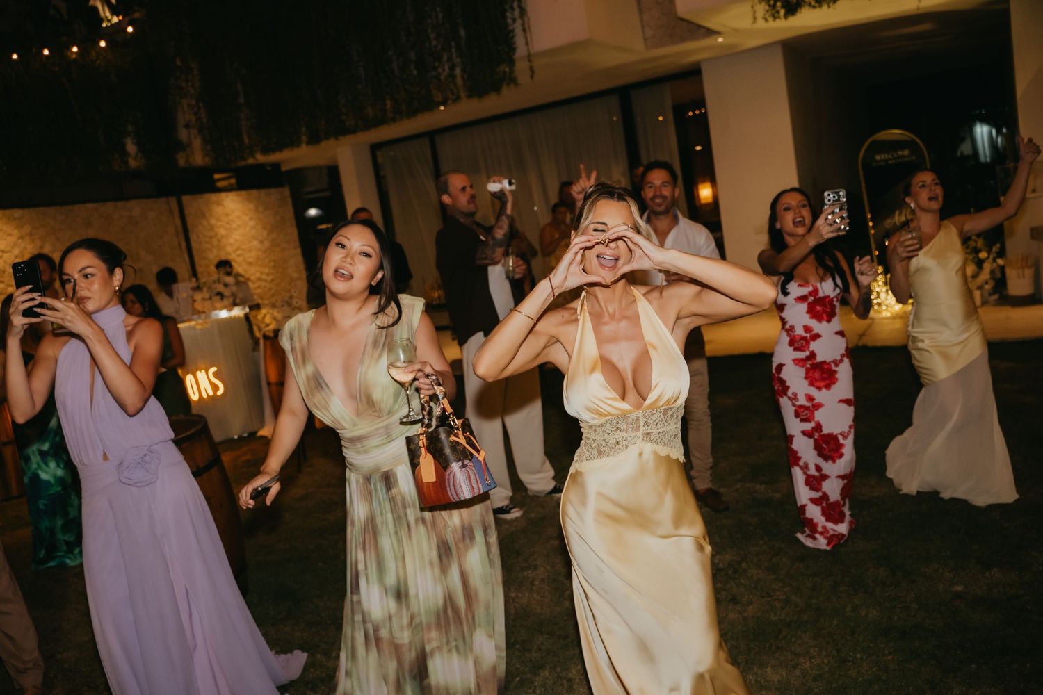 Bridesmaids in colorful gowns dance joyfully at lively outdoor tropical wedding reception at night.